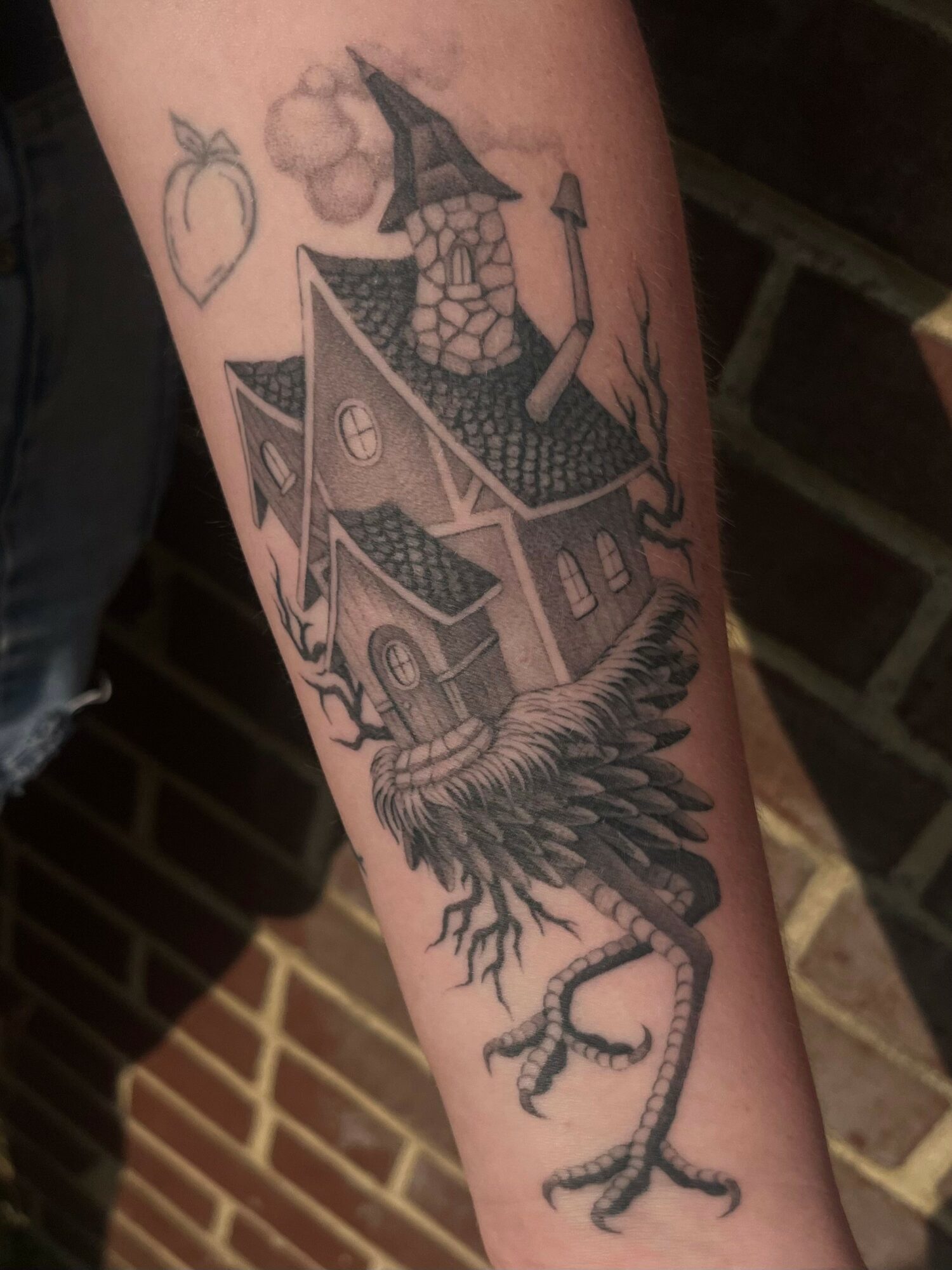 Tattoo of a house with a chimney, a tree, and a bird's legs with claws, on a person's arm.