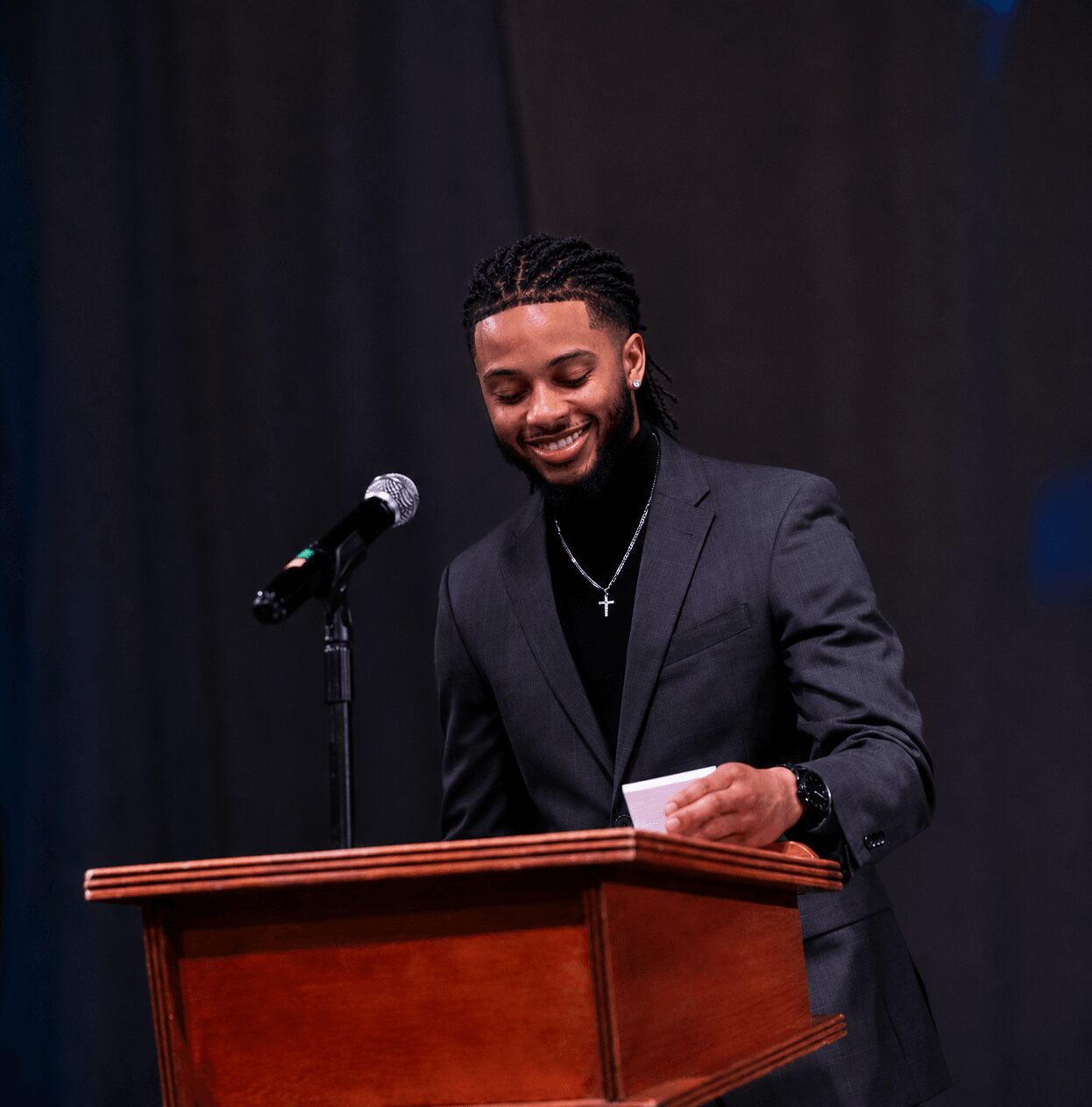 Man in a suit smiling at a podium with a microphone, holding a paper, against a dark background.