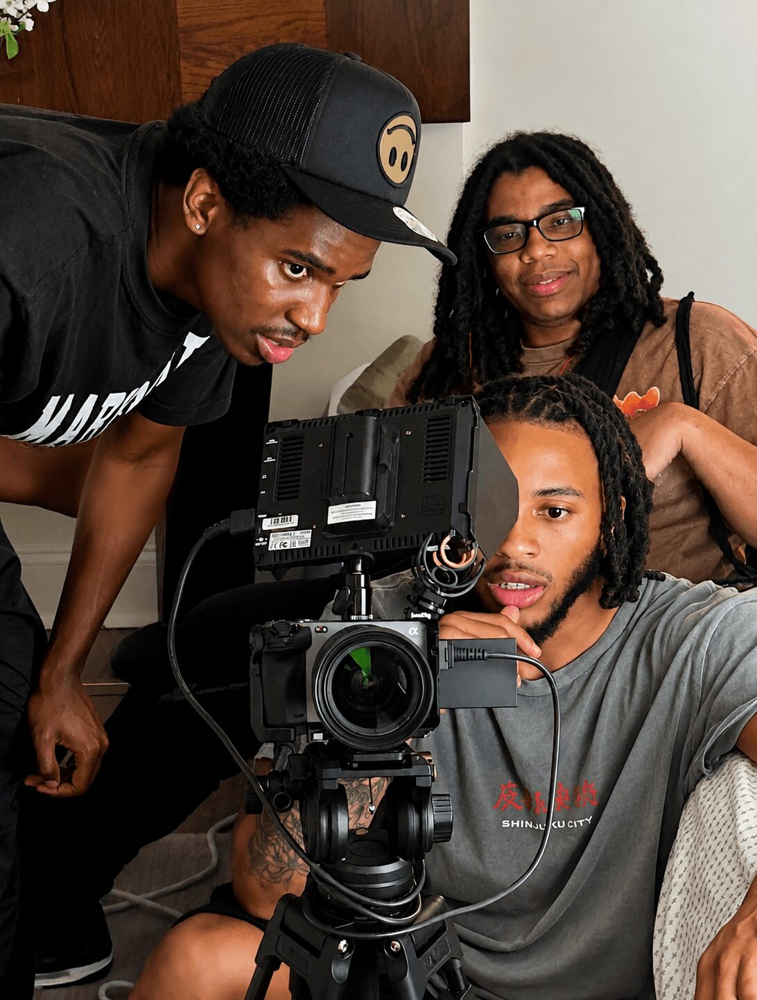 Three people gathered around a camera, one adjusting settings, two observing, indoors with a white wall background.