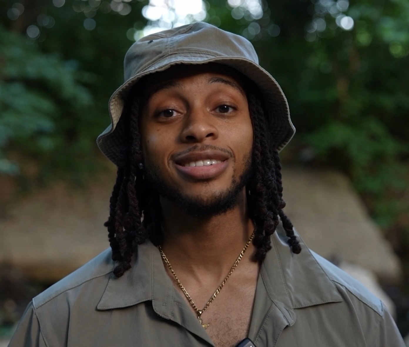 Young man outdoors wearing a bucket hat and gray shirt, smiling, with trees in the background.