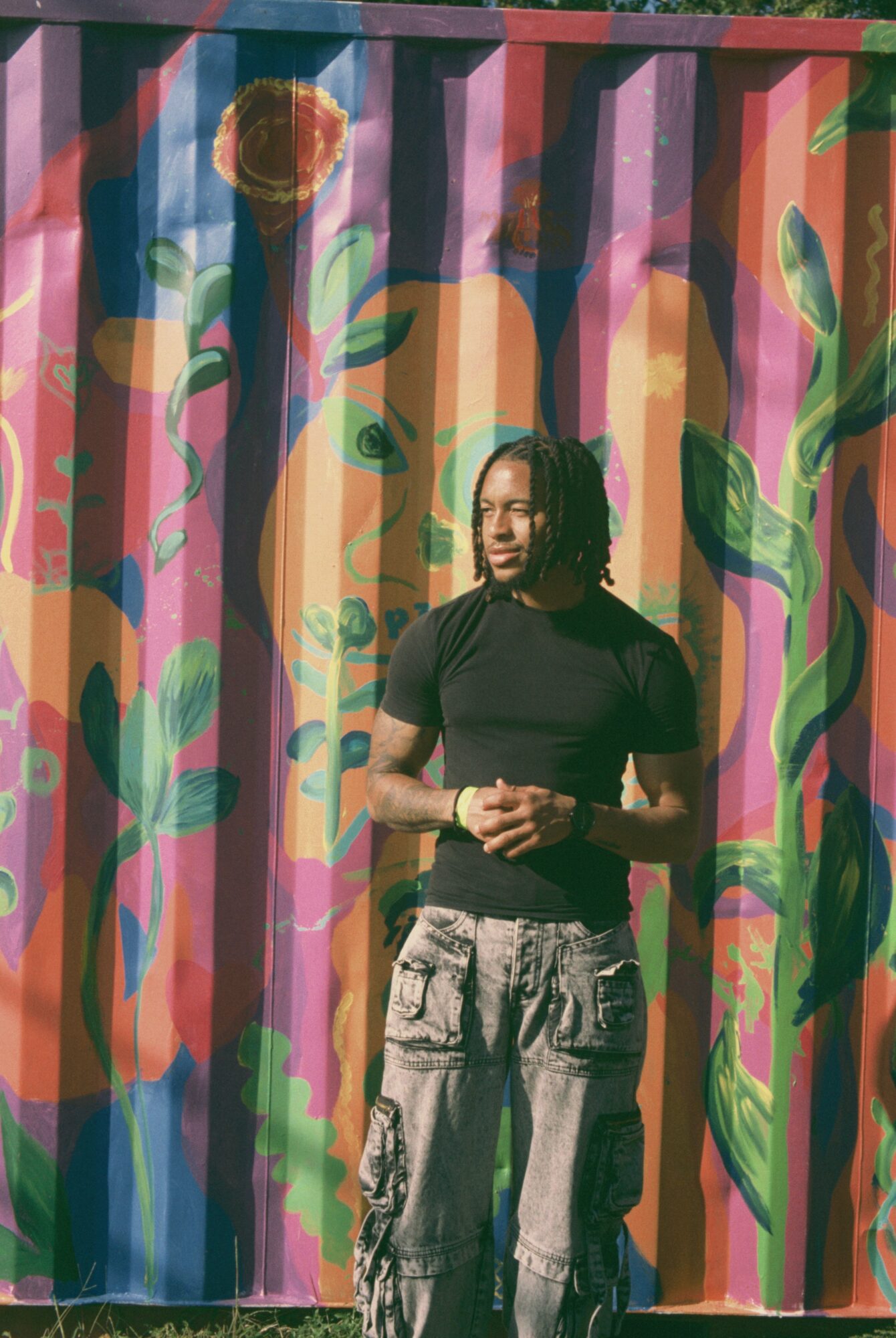 Person with curly hair wearing a black shirt and cargo pants stands in front of a colorful, painted metal wall.