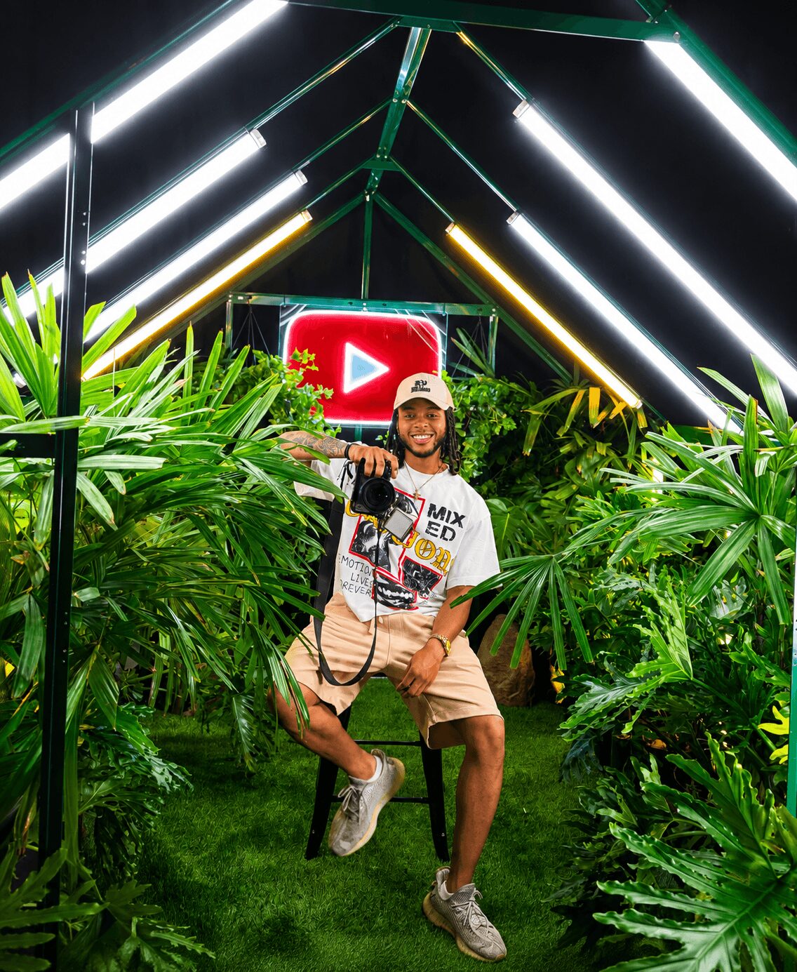 Person sitting on a swing surrounded by lush green plants, with a large illuminated YouTube logo in the background.