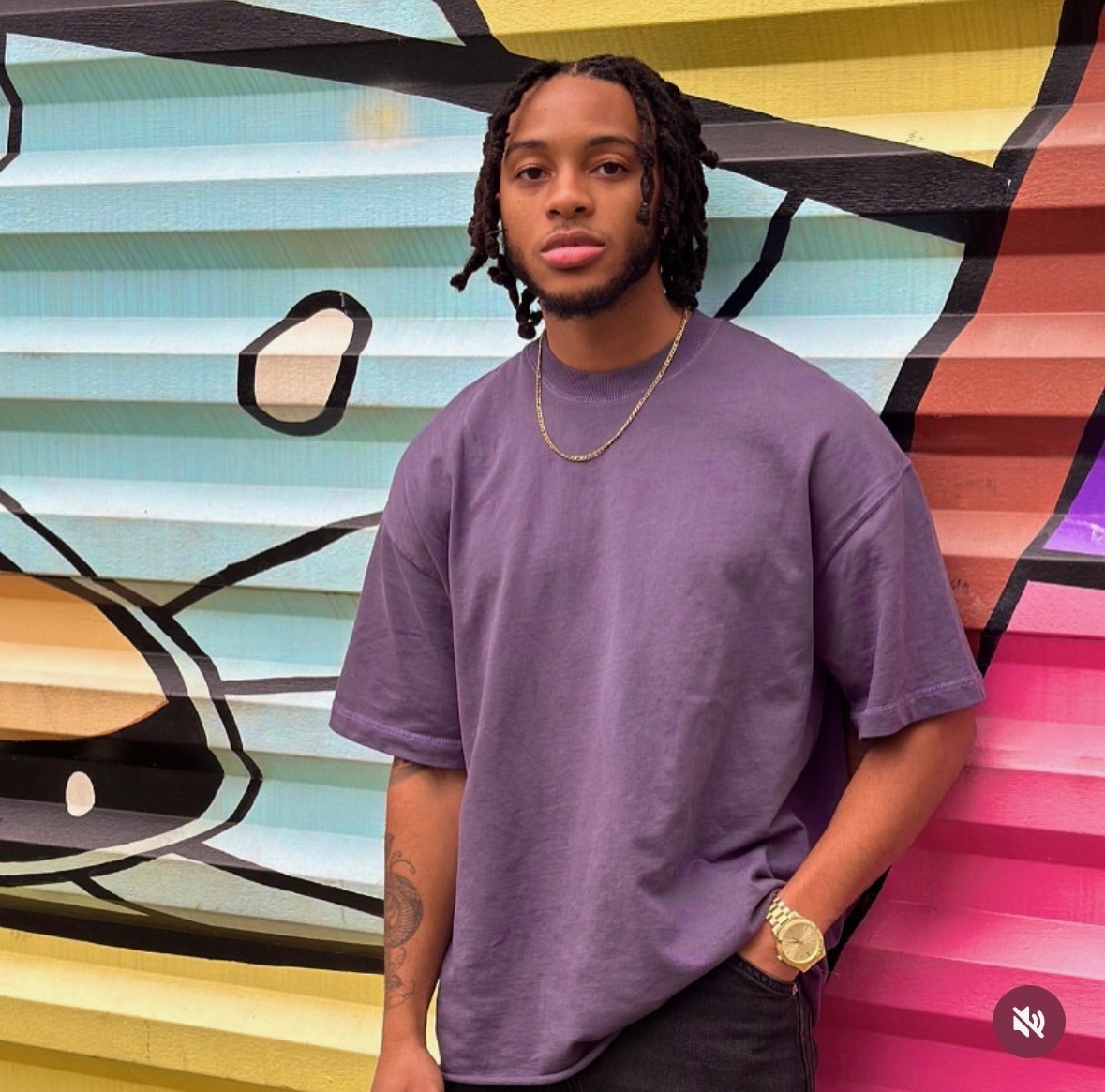 Young man with dreadlocks wearing a purple t-shirt and gold watch, standing in front of colorful graffiti wall.