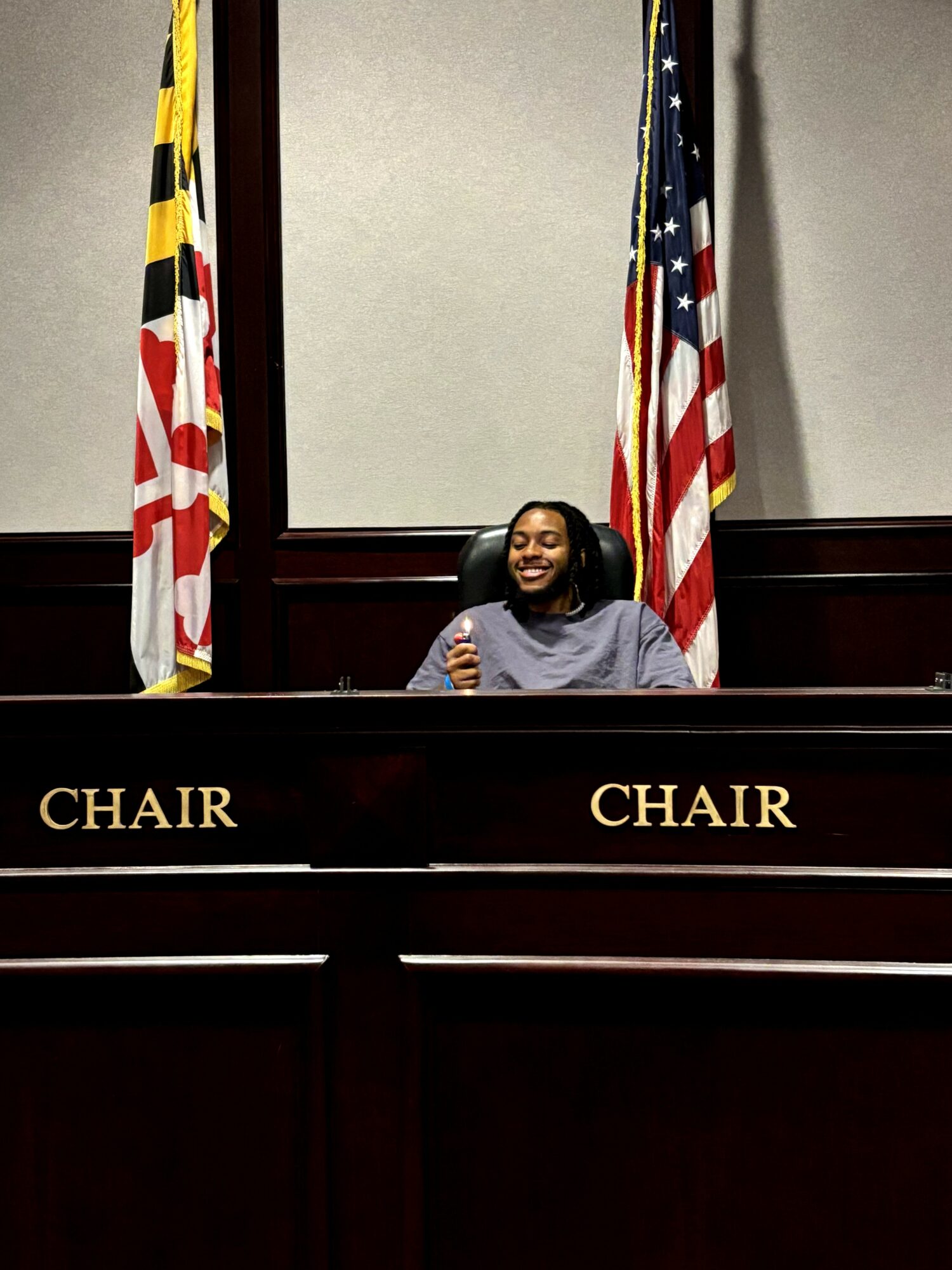 Woman sitting at a judge's bench with Maryland and U.S. flags behind her, smiling, in a courtroom setting.