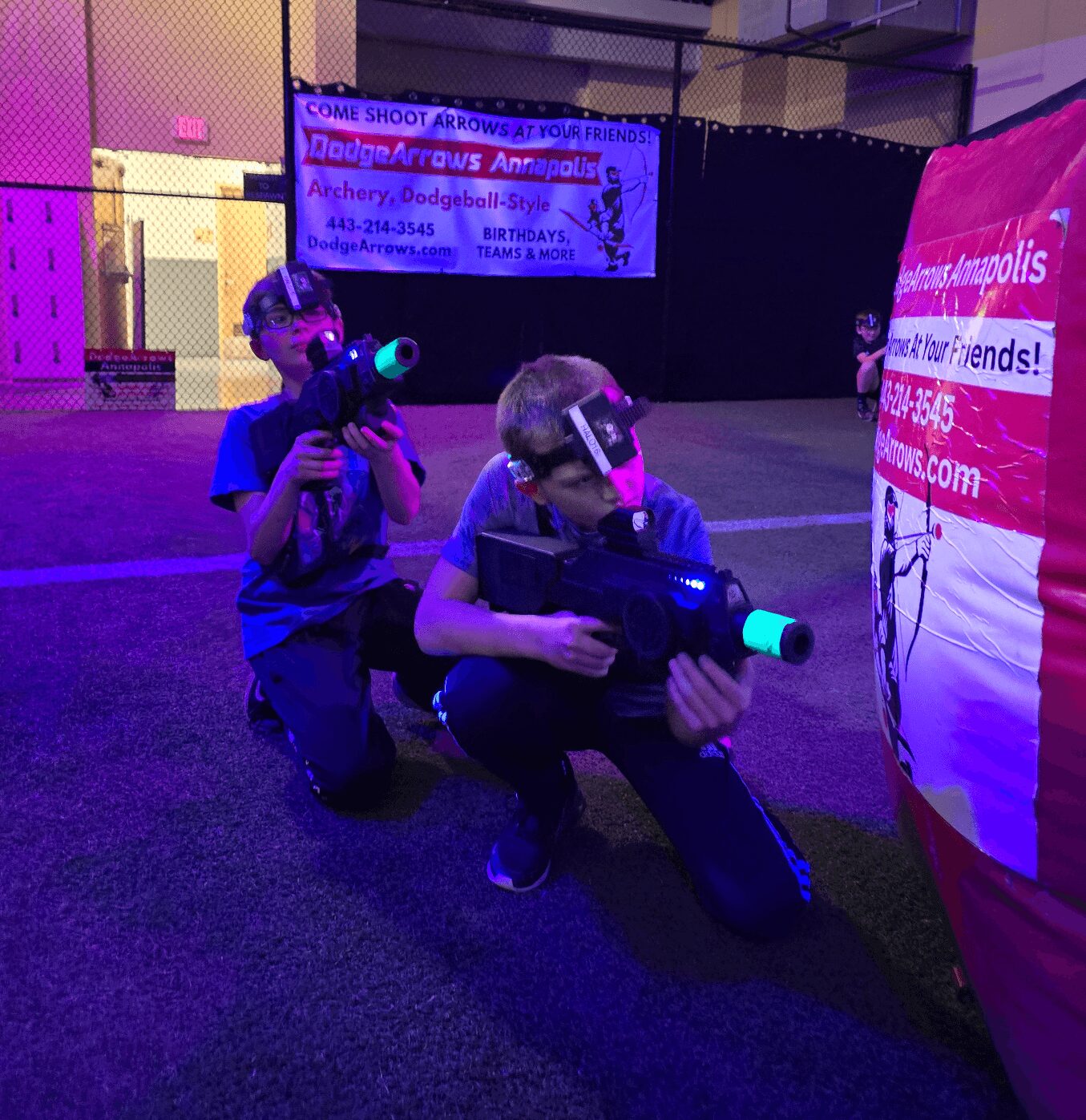 Two children wearing virtual reality headsets and holding laser tag guns indoors, kneeling on the floor. Bright lighting and banners in background.