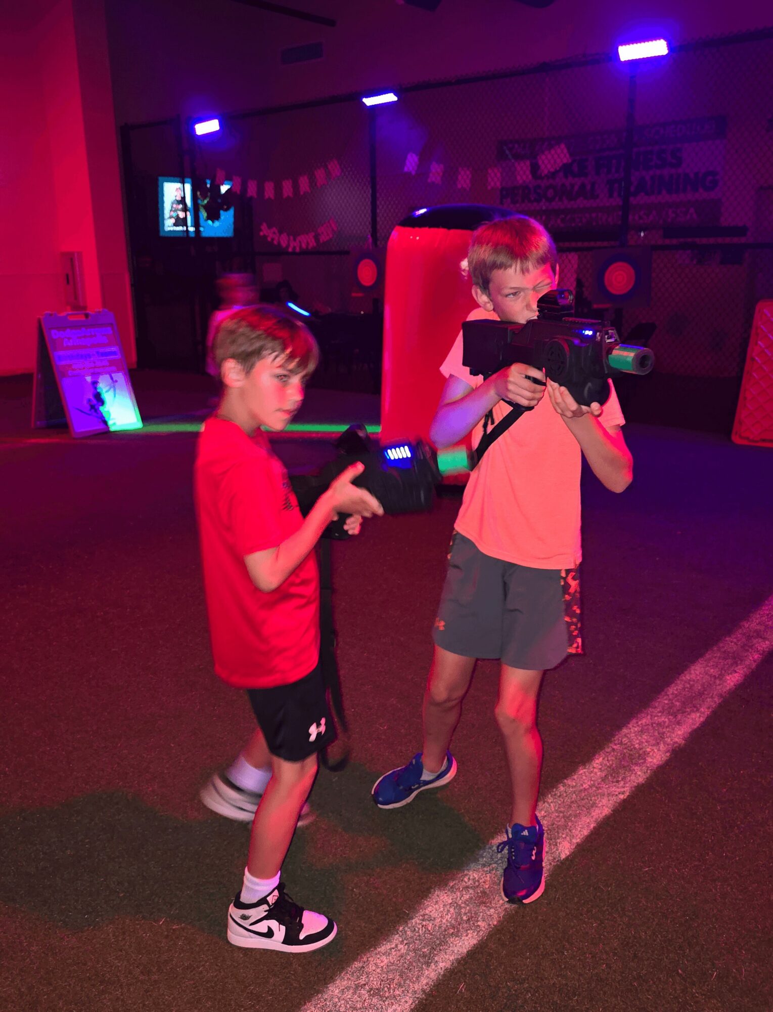Two children playing laser tag in a dark room with colorful lights, one holding a laser gun and the other pointing.