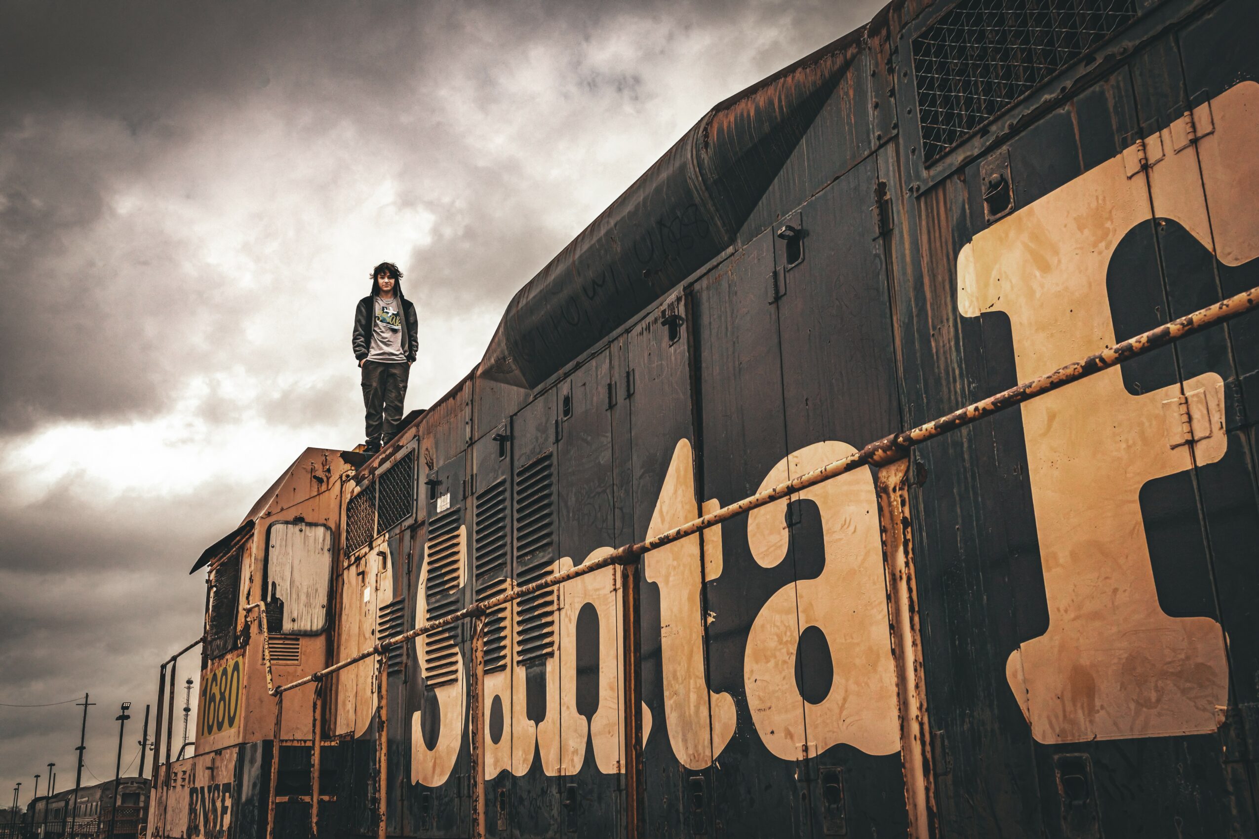 Person standing on a train with large graffiti letters on its side, cloudy sky in background.
