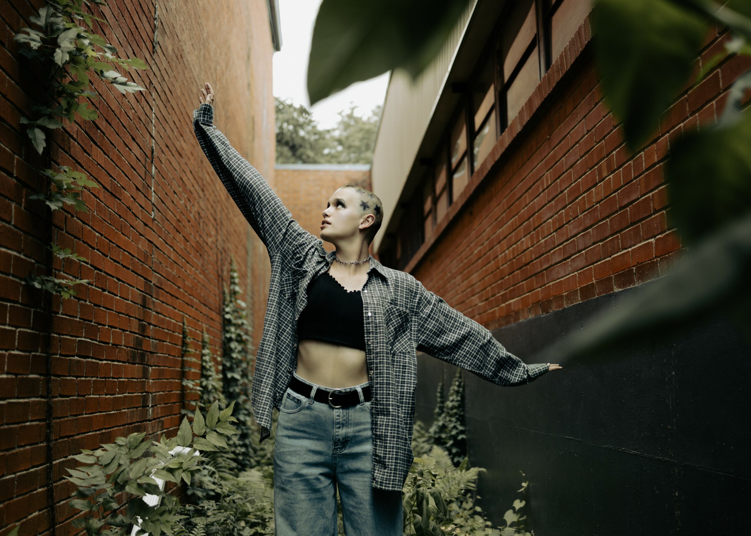 Person with short hair wearing a black crop top, plaid shirt, and jeans, standing between brick walls, reaching upward.