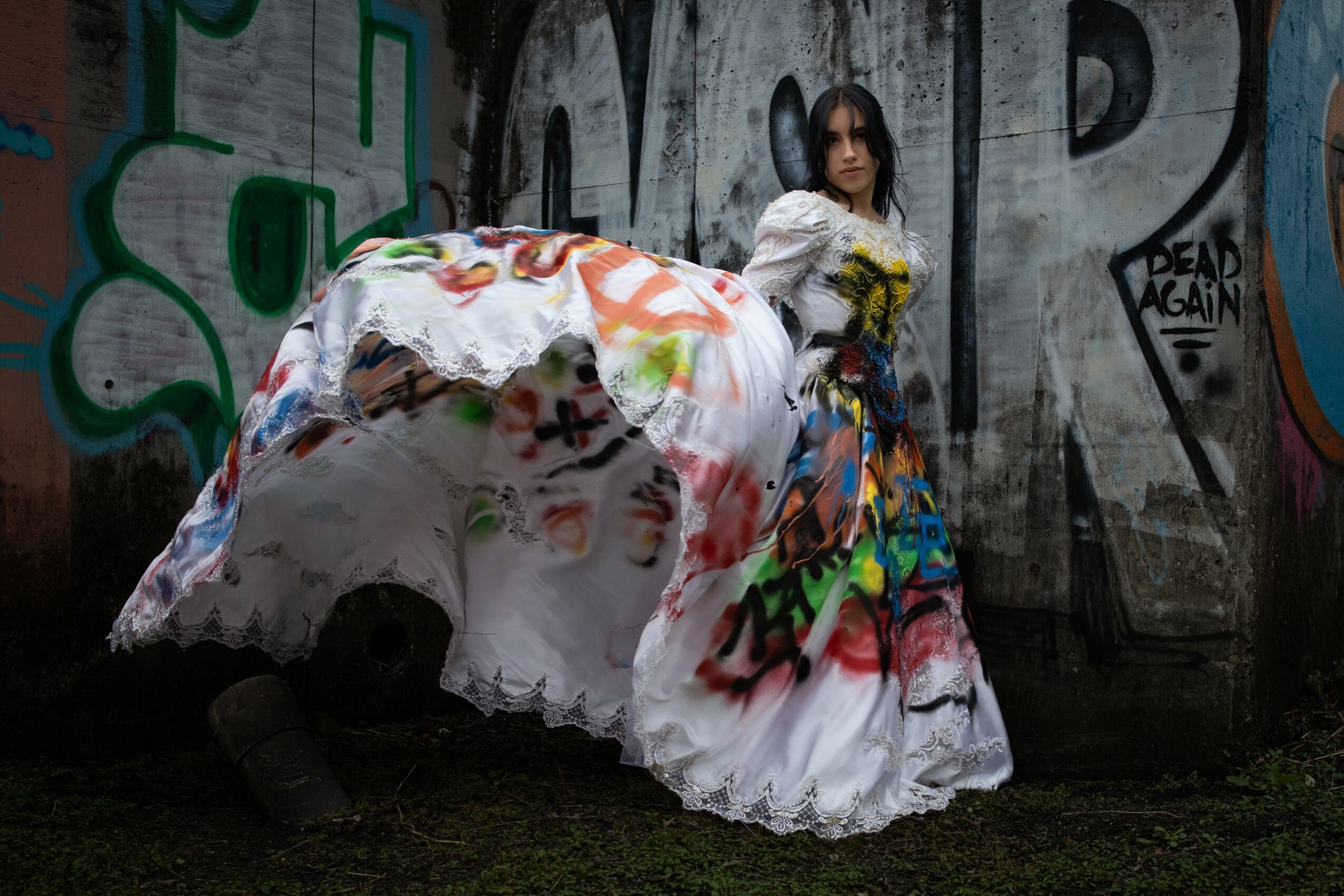 Person in a colorful, layered dress holding yellow flowers, standing in front of graffiti-covered wall.