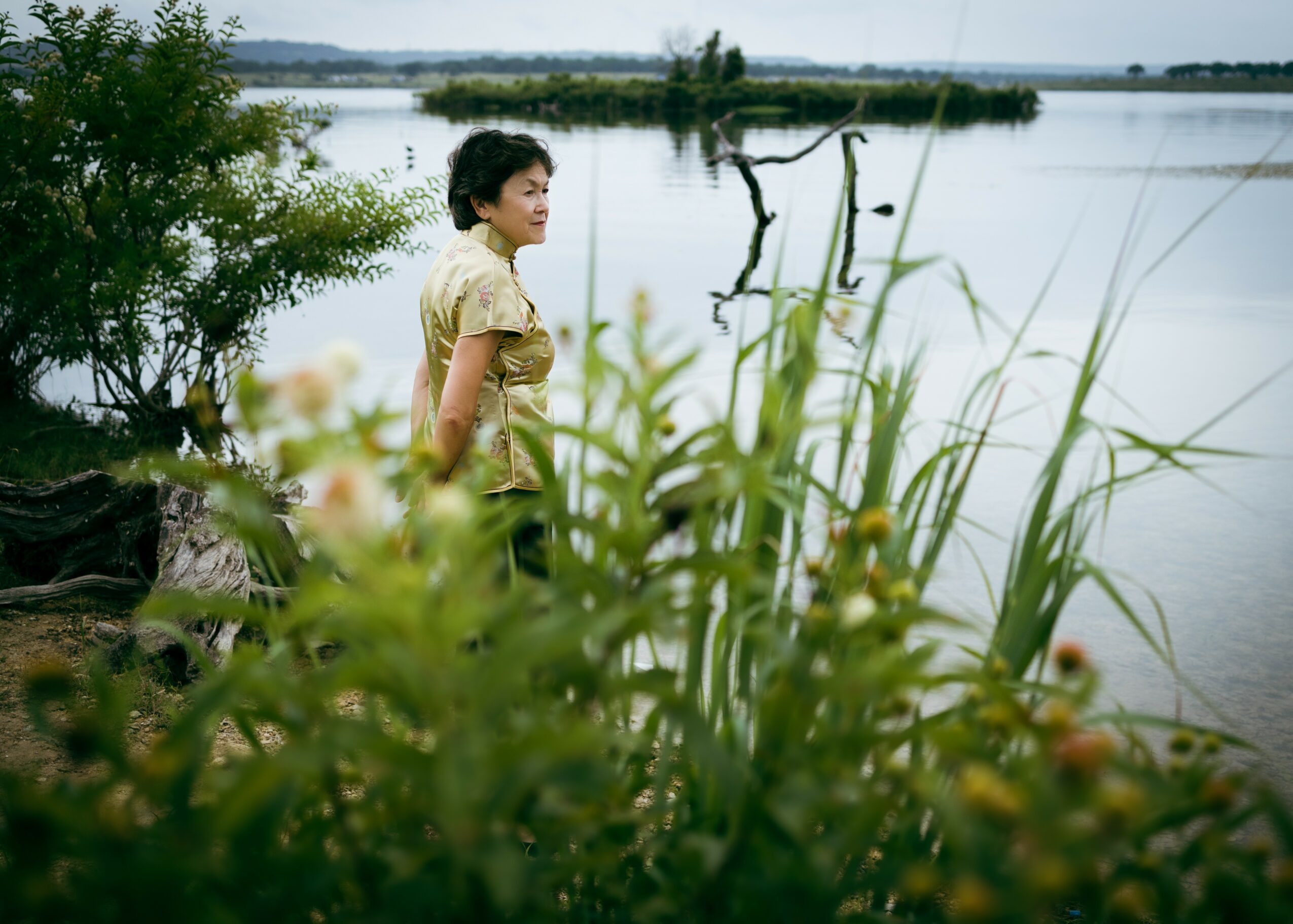 Woman standing near water with tall grass and trees in the background, looking to the side.