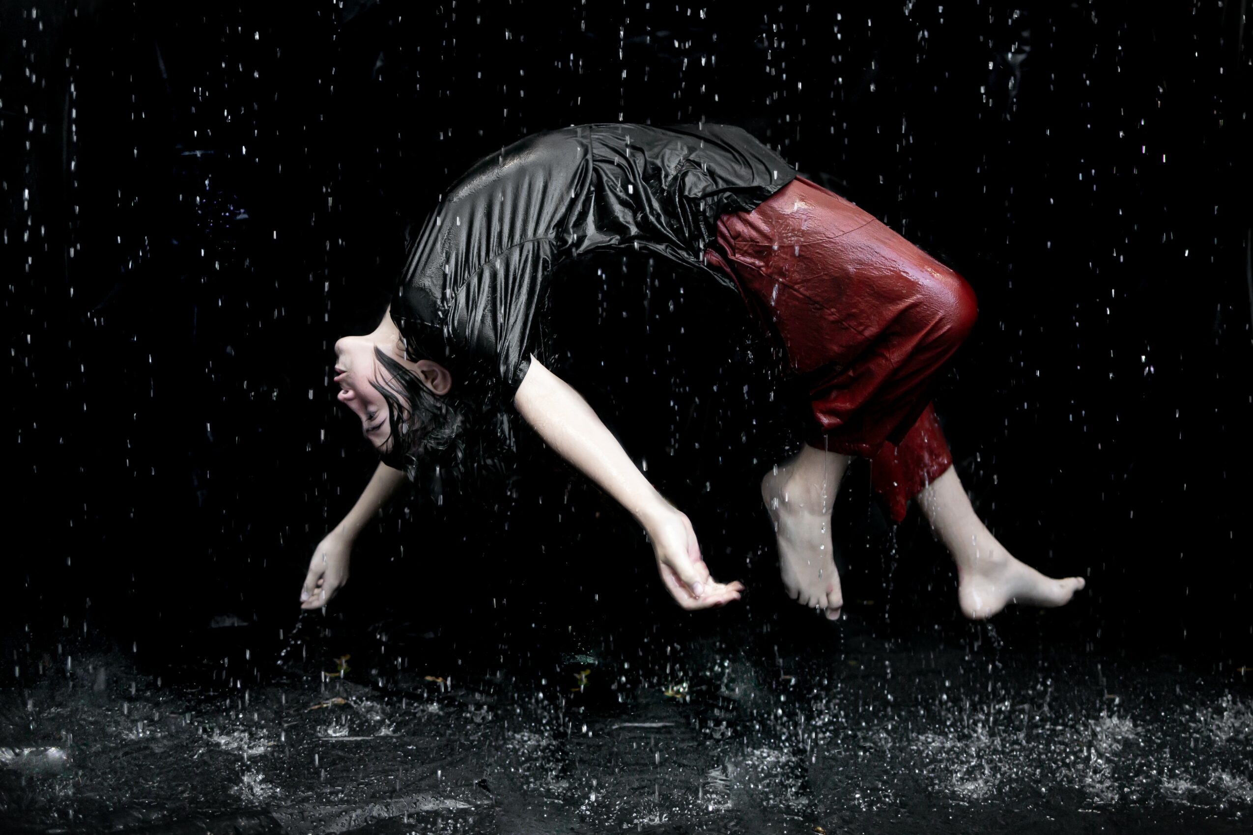 Person performing a backbend on stage with water splashing around, wearing a black top and red pants.