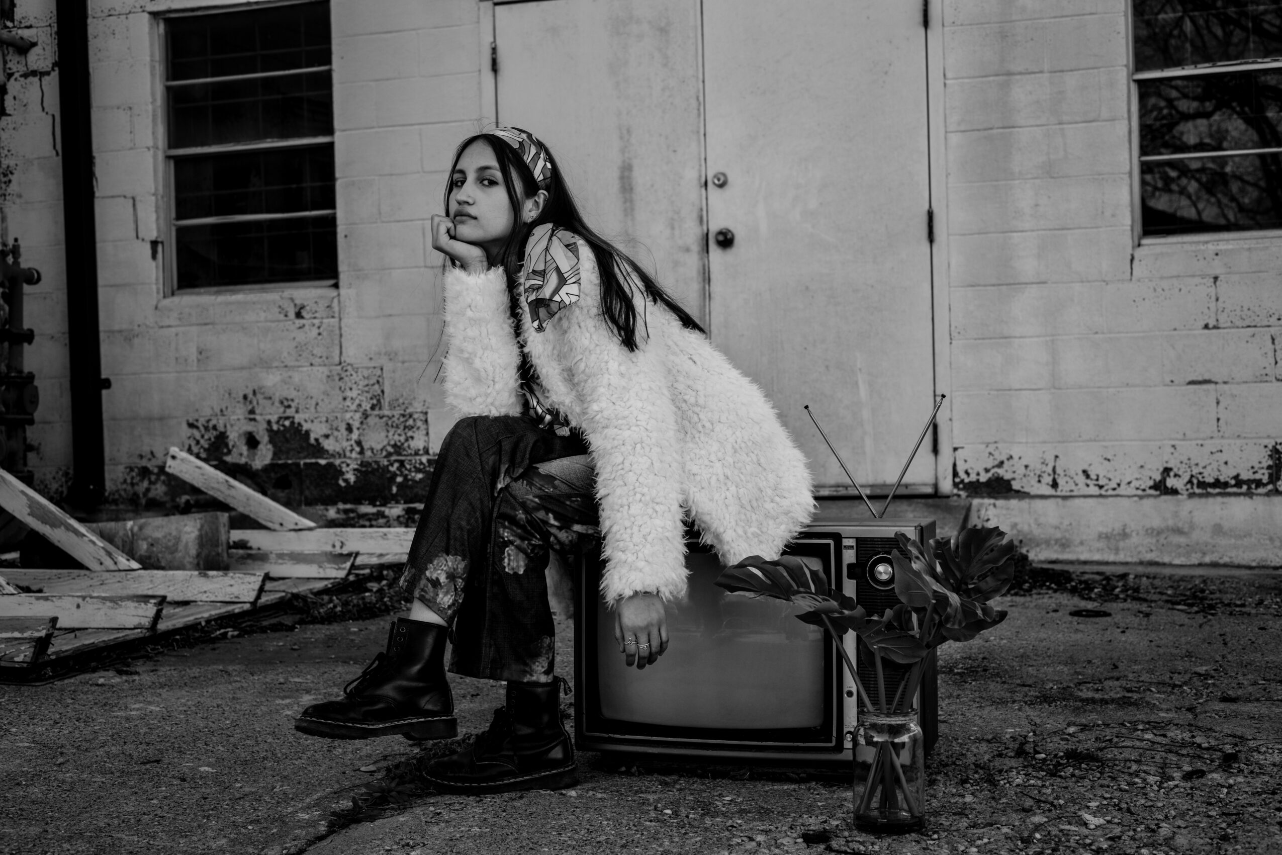 A woman sits on a vintage television outside a weathered building, resting her chin on her hand, in black and white.