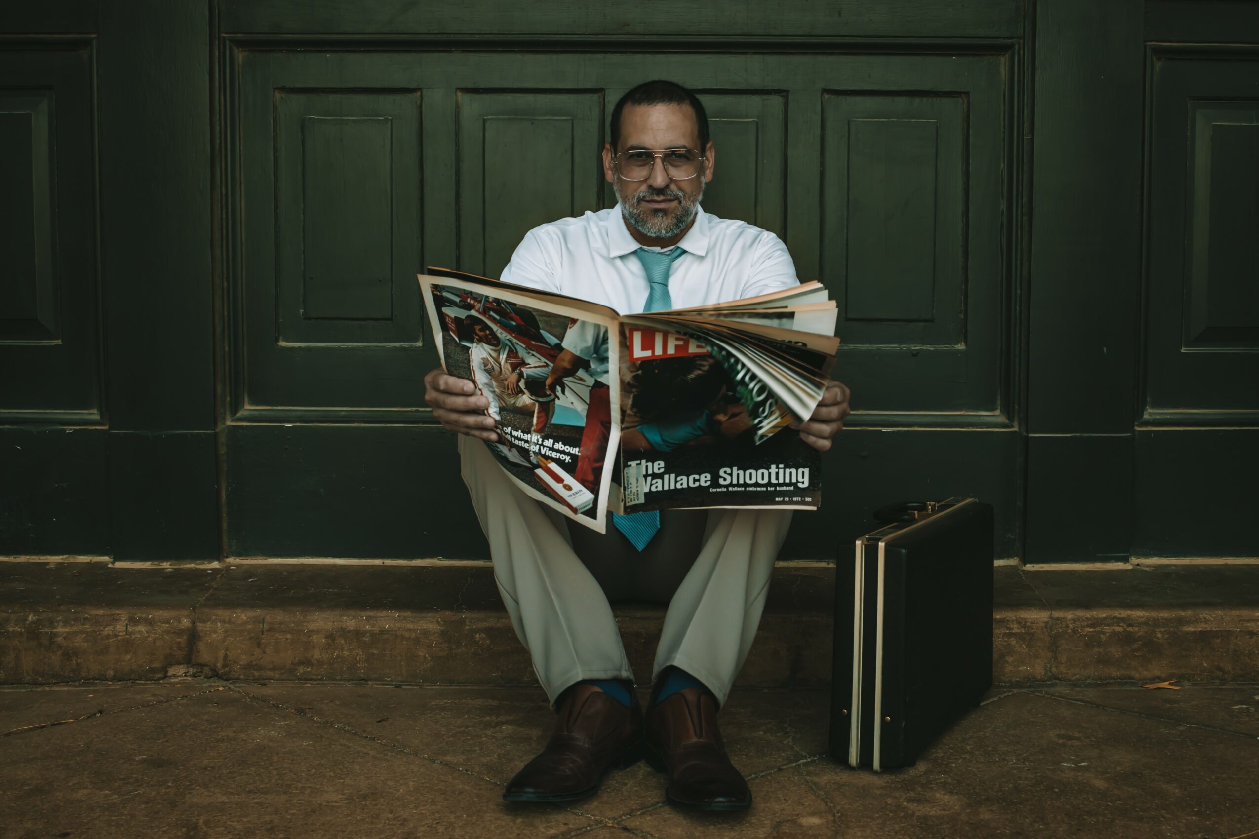 Man sitting on the floor reading a newspaper, with a briefcase beside him, against a green paneled wall.