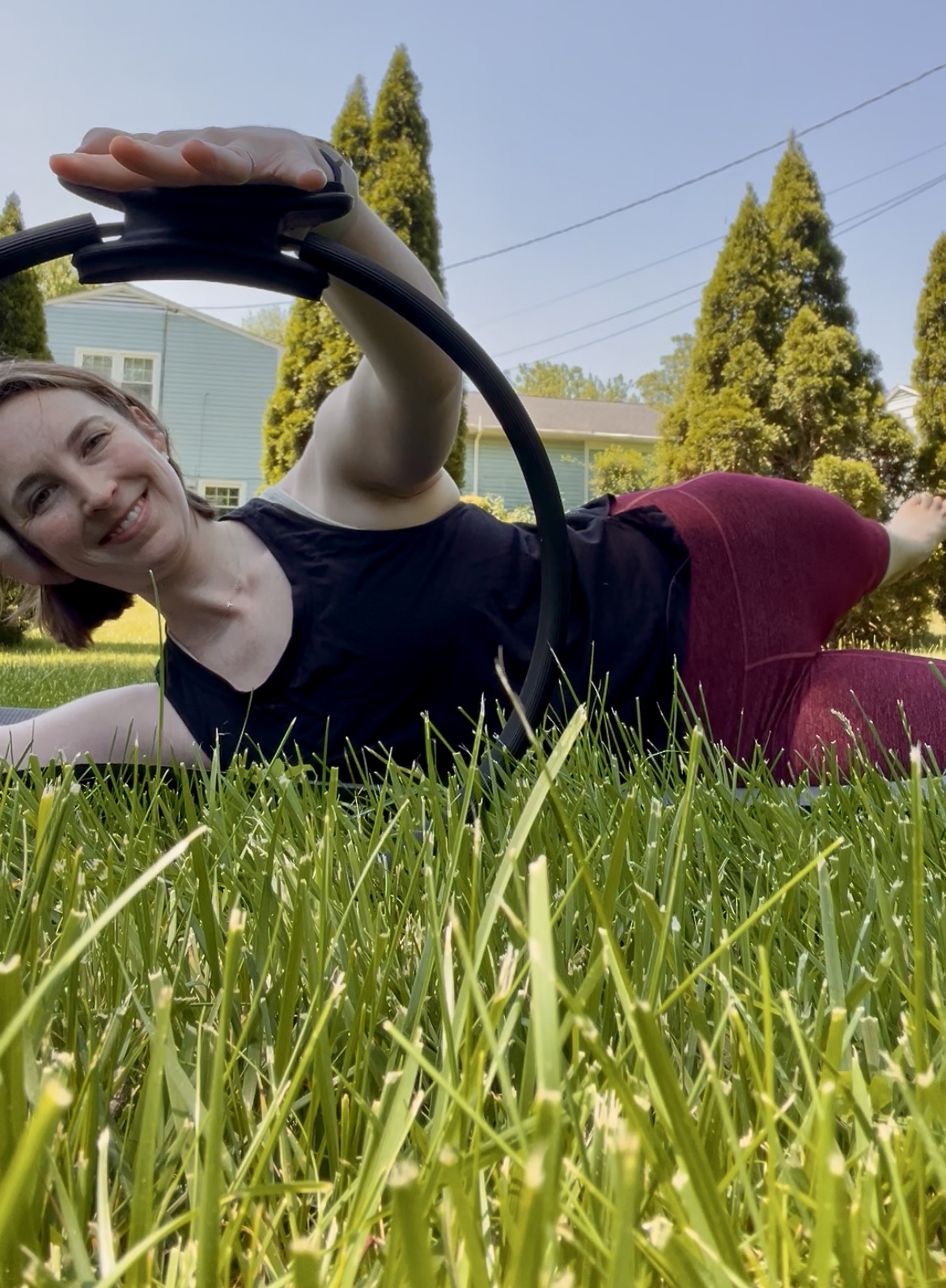 Young woman lying on grass outdoors, smiling, holding a black circular object, with trees and houses in background.