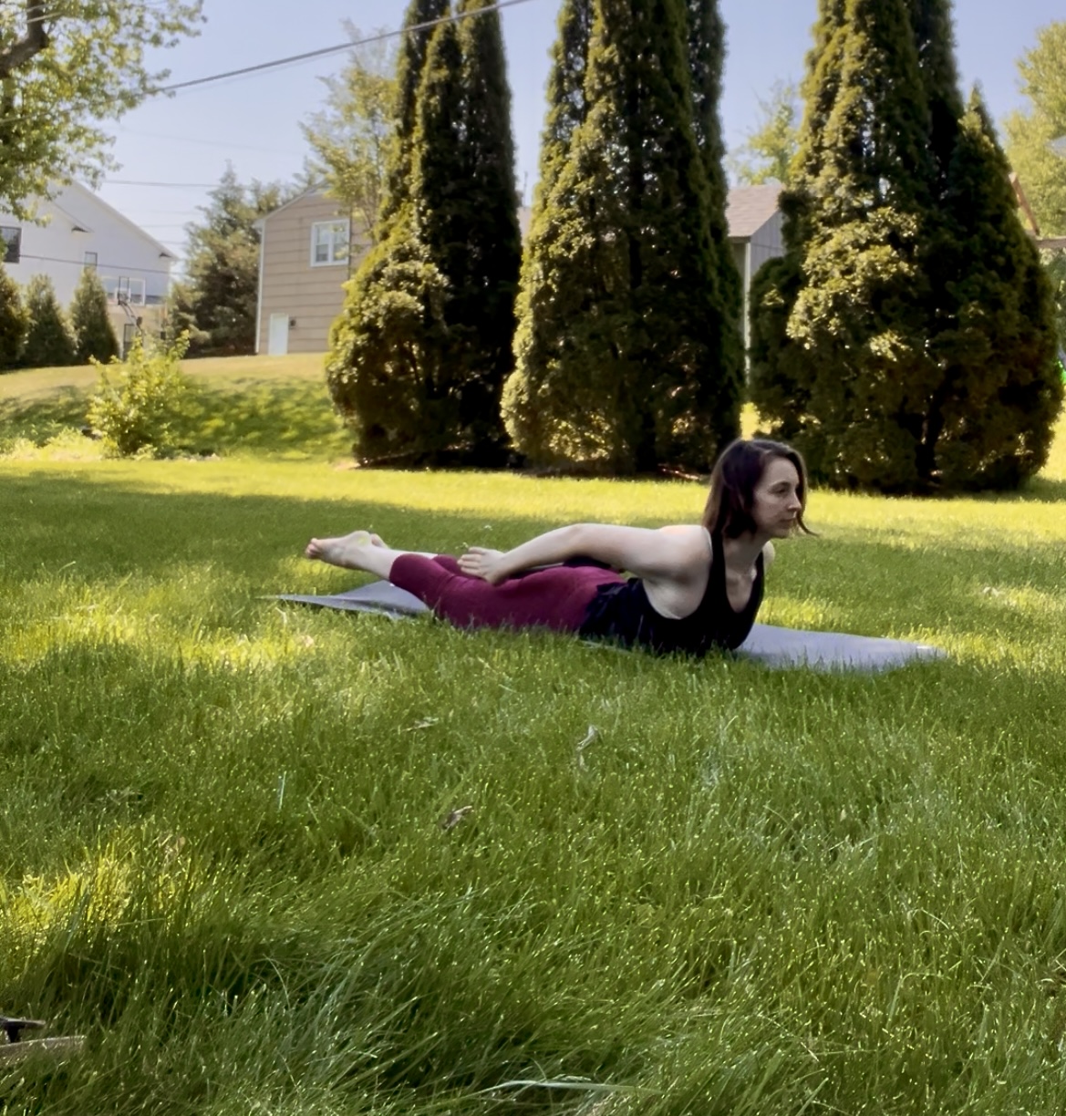 Woman practicing yoga outdoors on grass, lying on her stomach with arms extended forward, trees and houses in background.