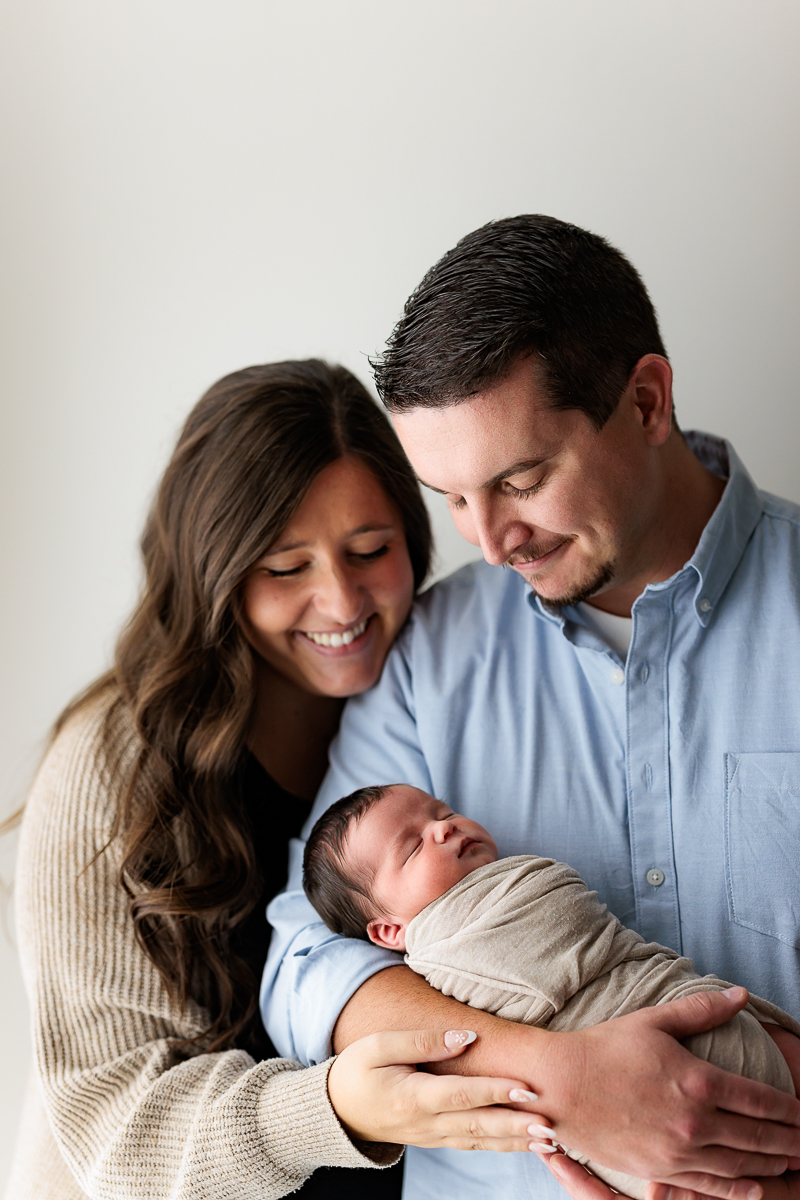 A family of three, a woman, a man, and a newborn baby, smiling and looking at the baby, against a plain background.