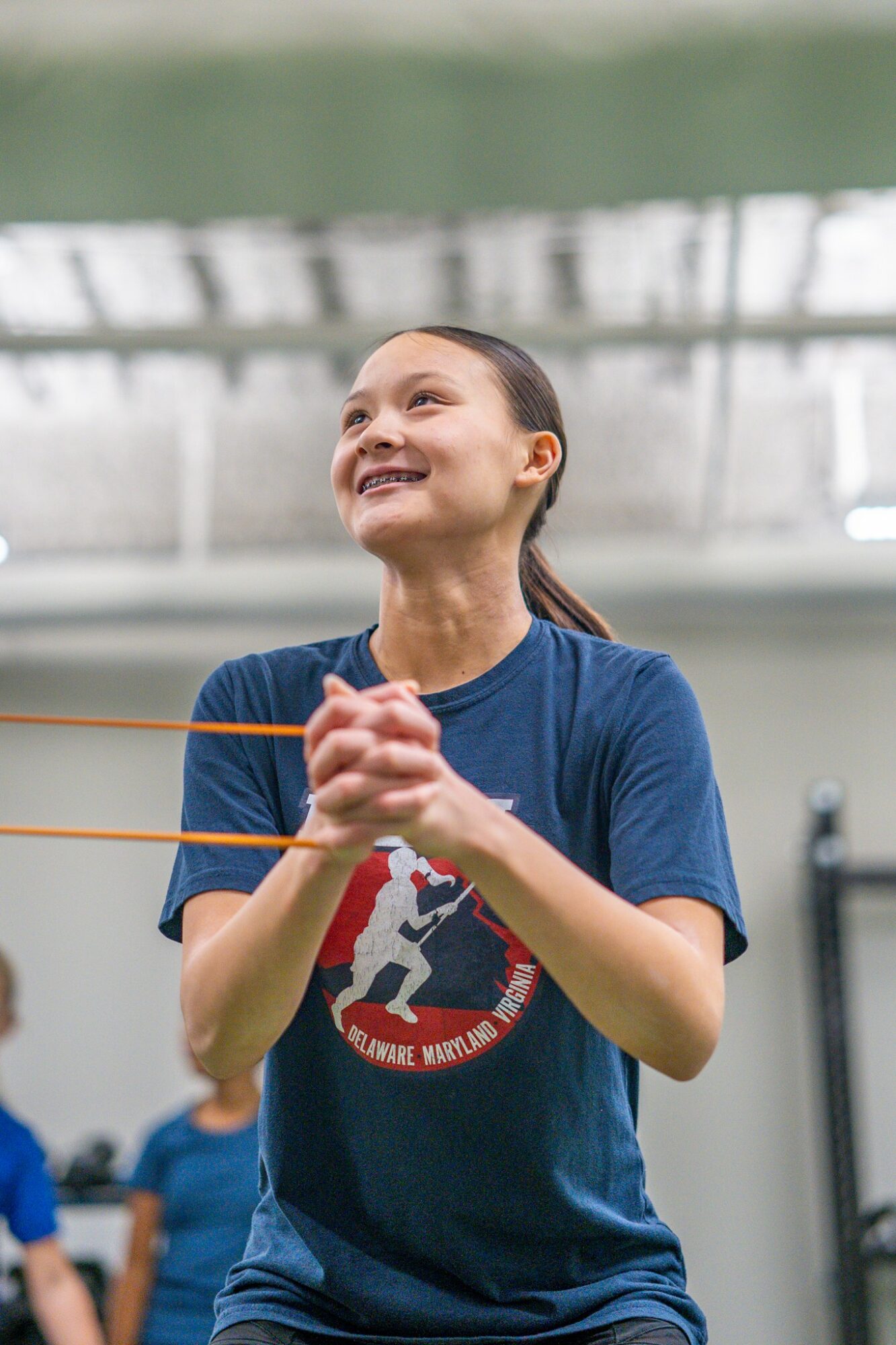Young girl smiling and clapping indoors, wearing a blue t-shirt with a logo, with a blurred background.
