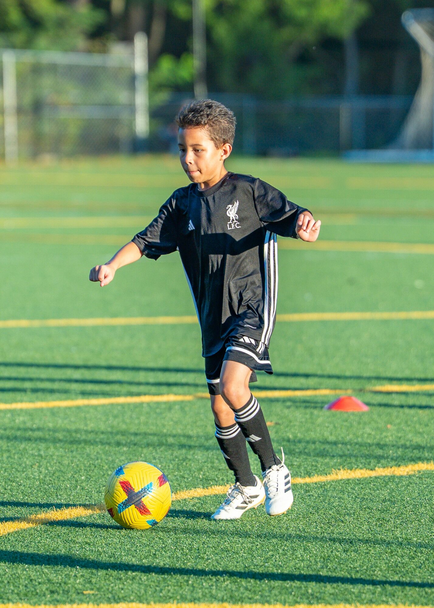 Child playing soccer on a field, wearing a black sports outfit, kicking a yellow ball with a red and blue design.