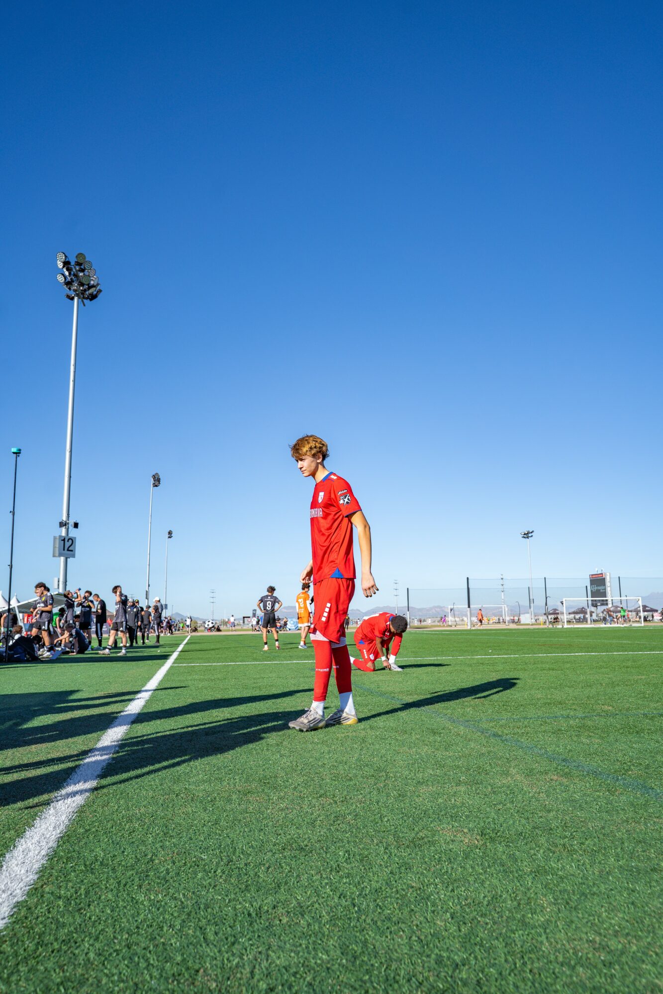 A soccer player in a red uniform stands on a green field with a clear blue sky background.