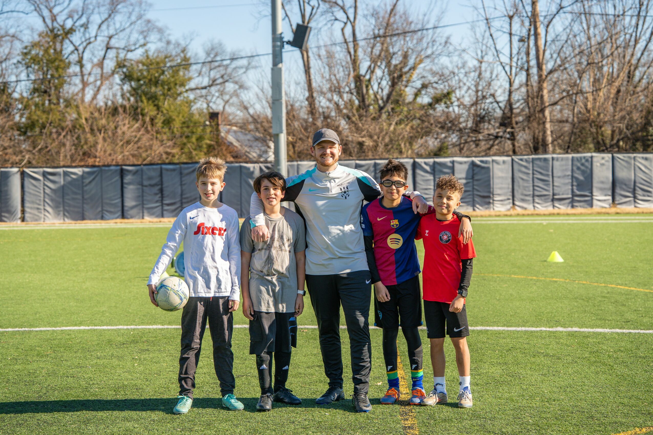 Group of five children and an adult standing on a soccer field, with trees and a fence in the background.