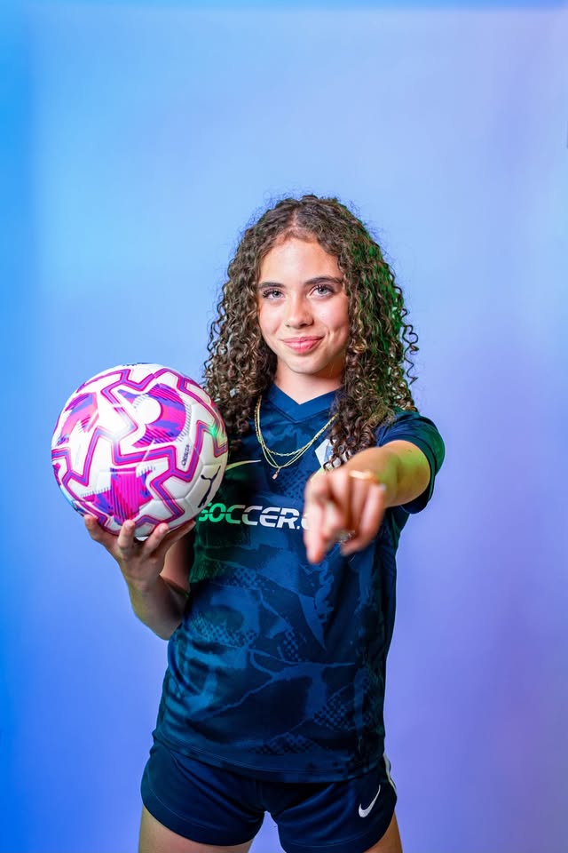 Young girl with curly hair holding a pink and white soccer ball, pointing forward, smiling, wearing a dark sports jersey.