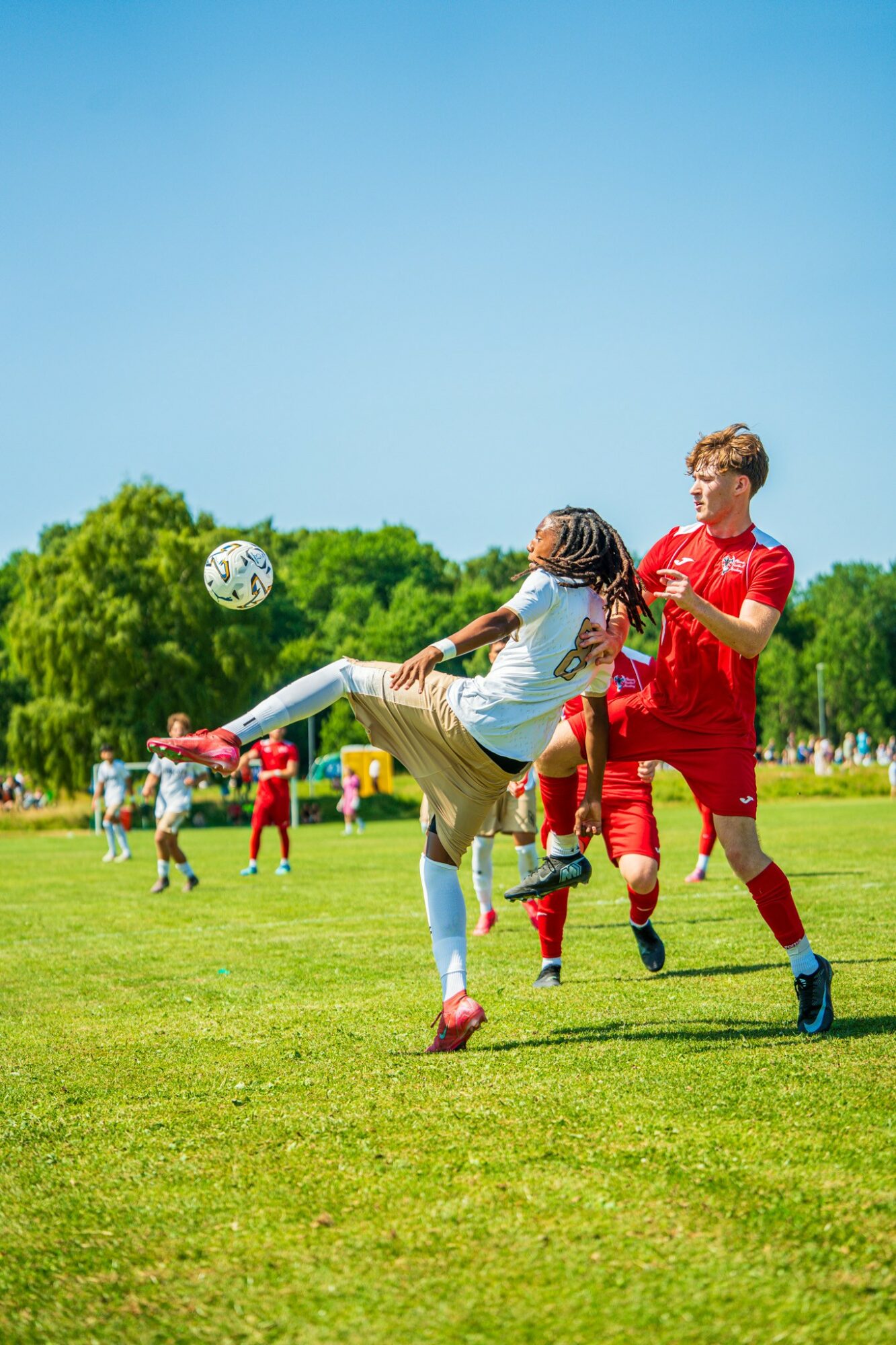 Children playing soccer on a grassy field under a clear blue sky, with trees in the background.