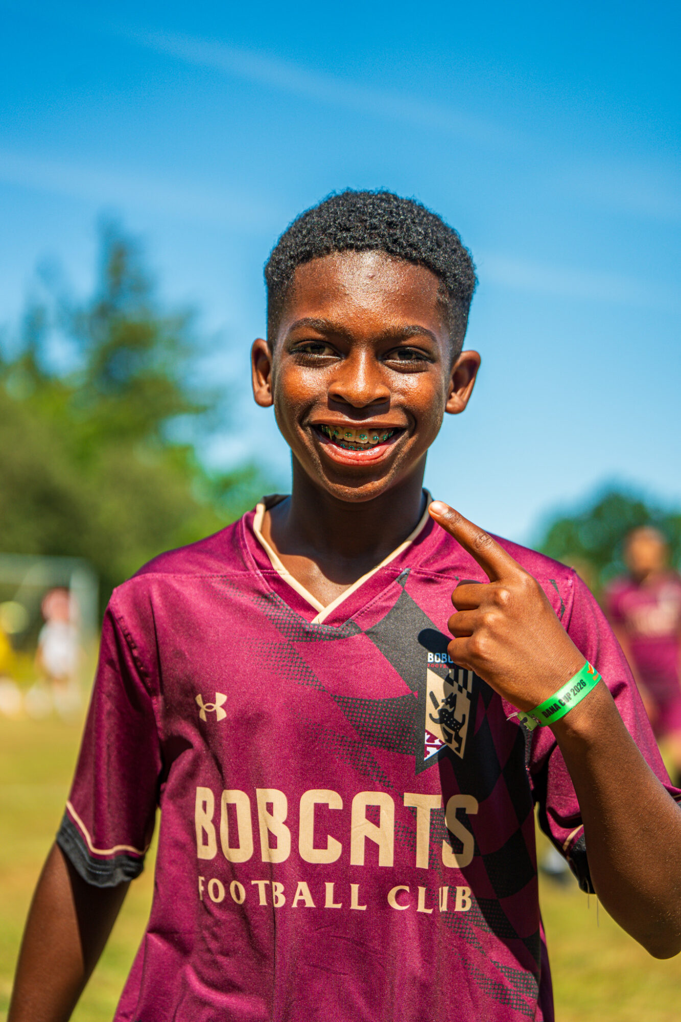 Young person smiling, wearing a maroon football jersey, pointing to their face outdoors on a sunny day.