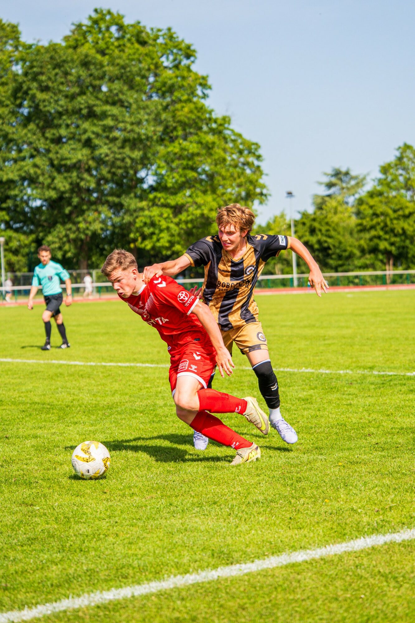 Two soccer players compete for ball on field, one in red, one in black and yellow, with trees and sky in background.