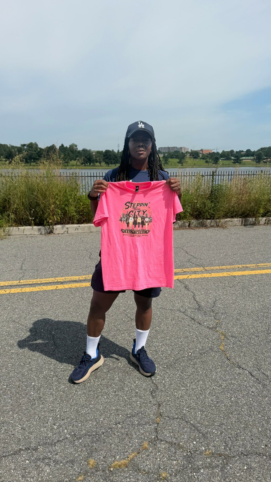 Person holding a pink T-shirt outdoors on a paved road with grass and water in the background.