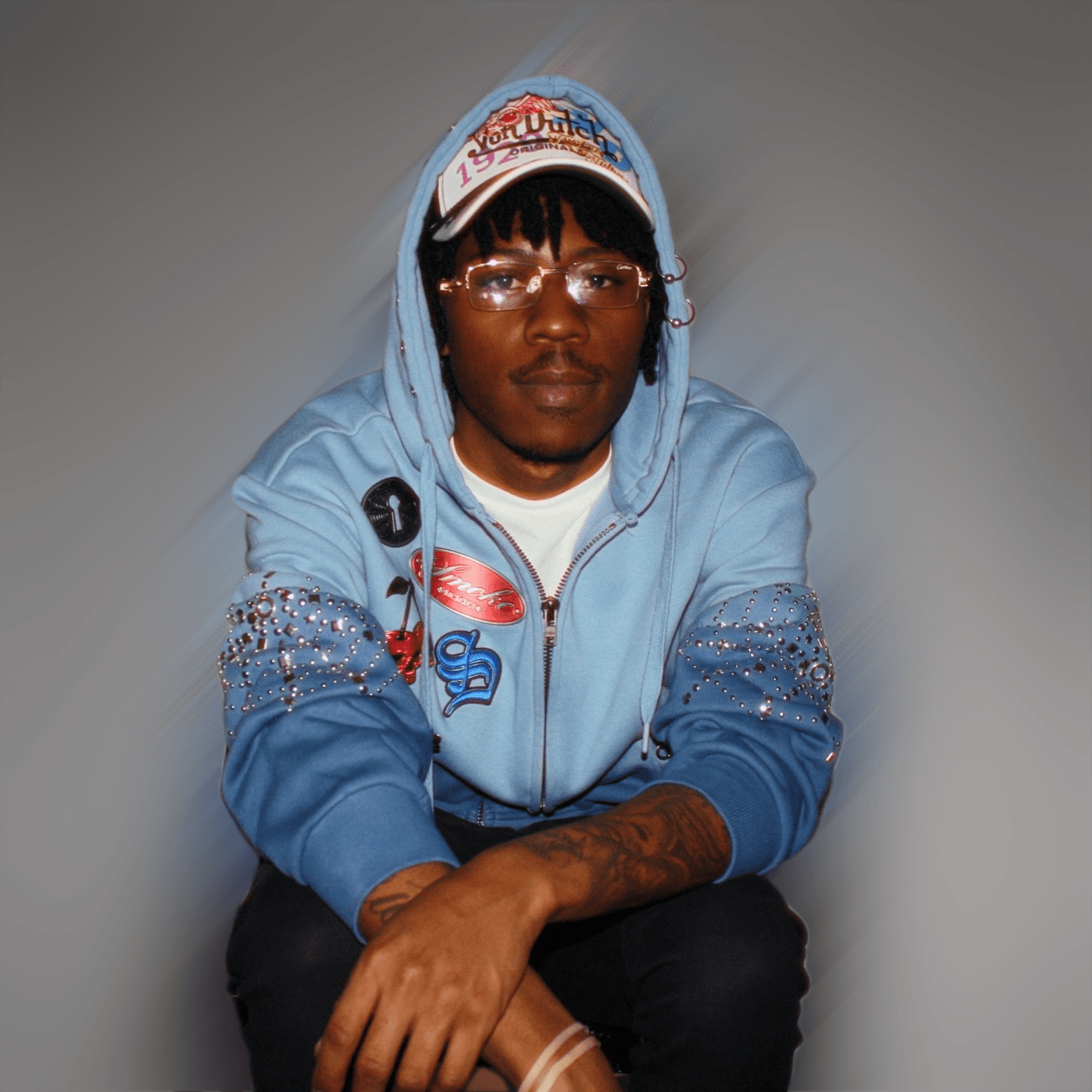 Young person wearing glasses, a light blue hoodie with patches, and a cap, sitting with arms crossed.