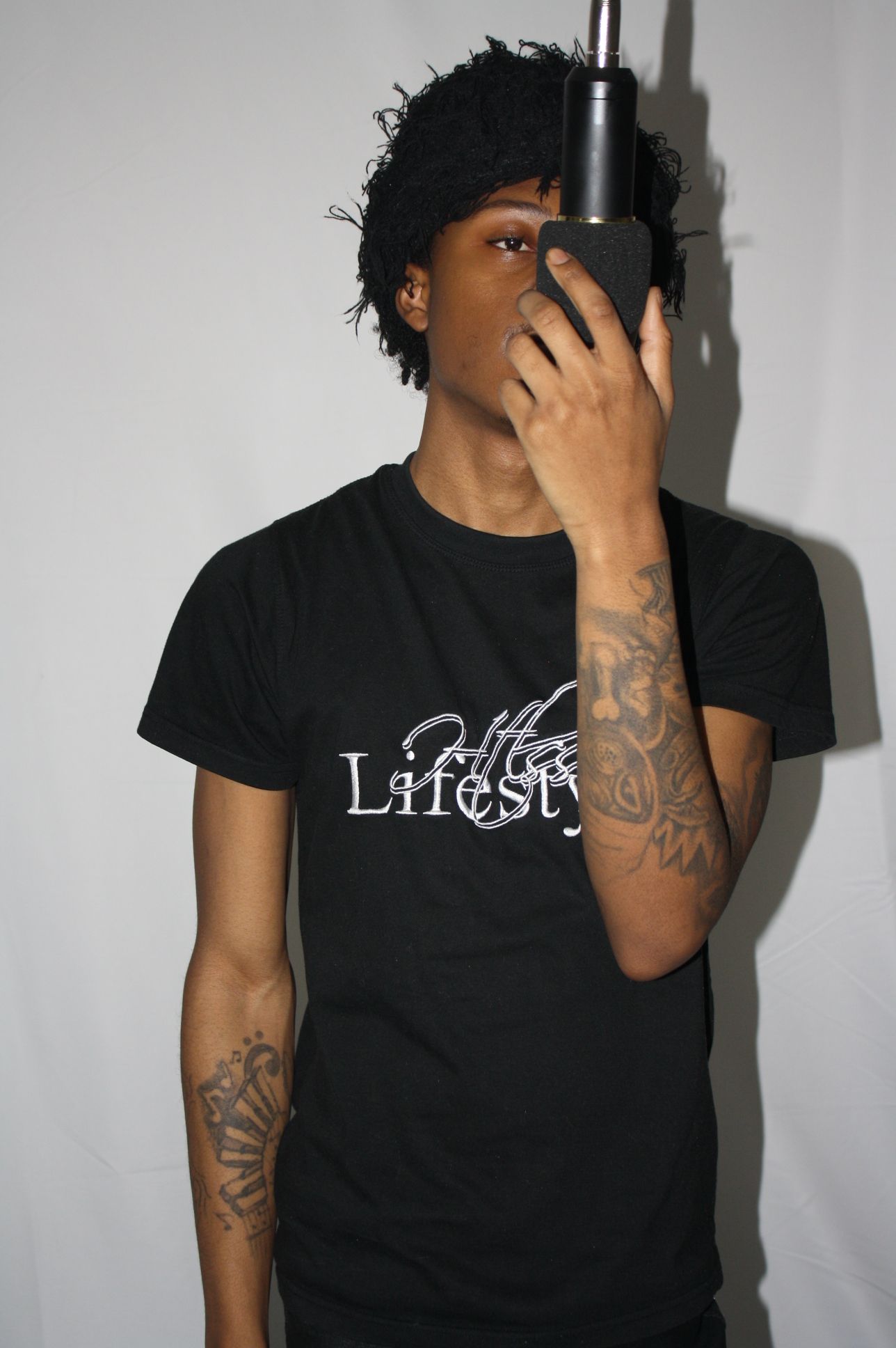 Person lying sideways holding a black object to their ear, with tattoos on their arm, against a plain background.