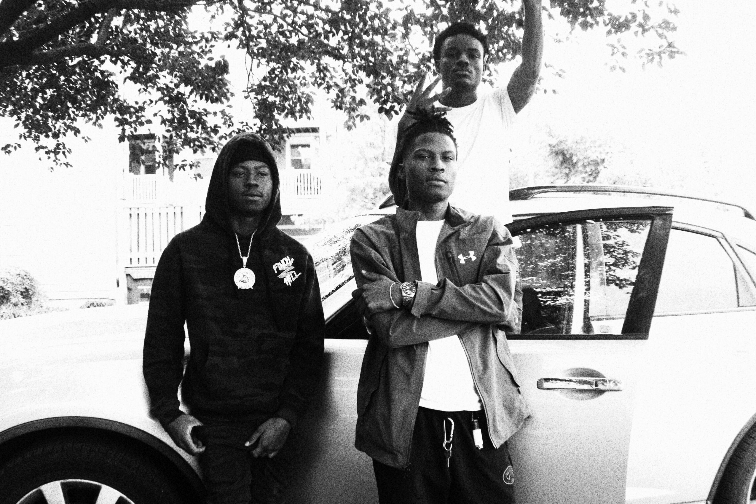 Three people pose outdoors near a car, with trees and a fence in the background, in black and white.