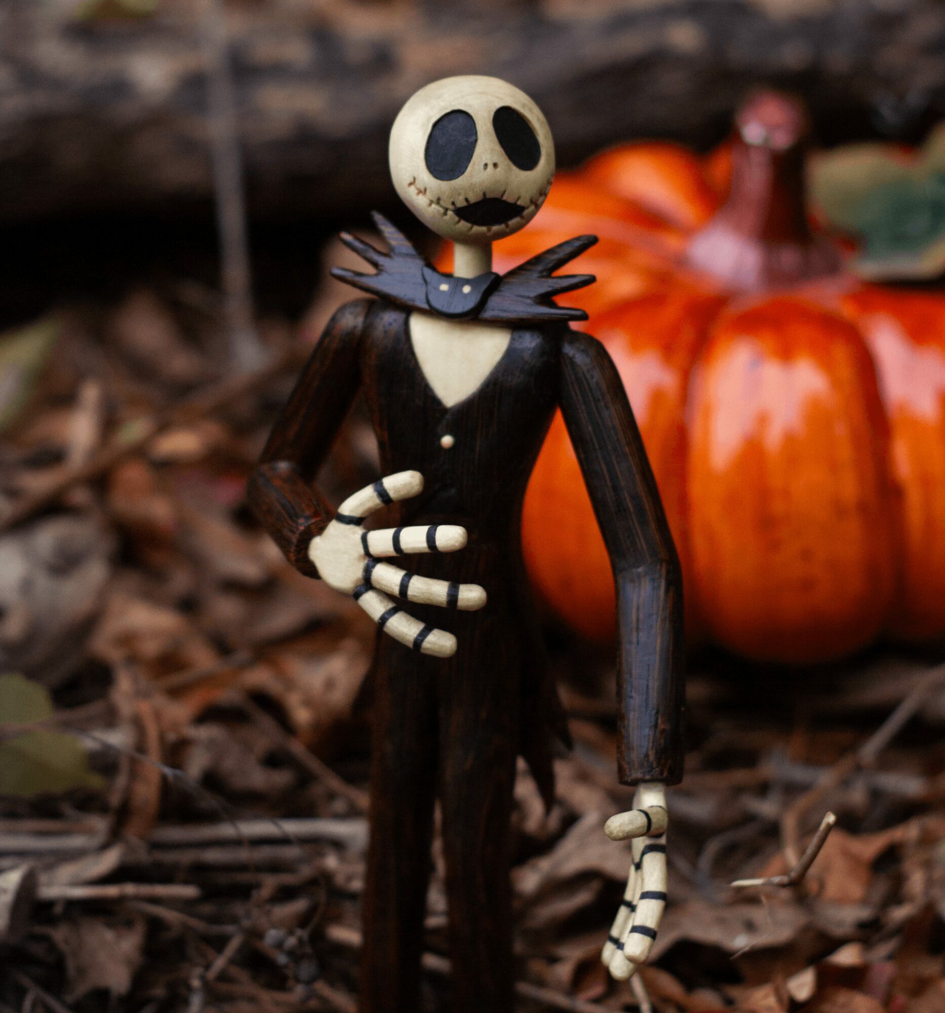 A stylized skeleton figure with a round skull, black eye sockets, and a black outfit, standing outdoors with pumpkins and fallen leaves.
