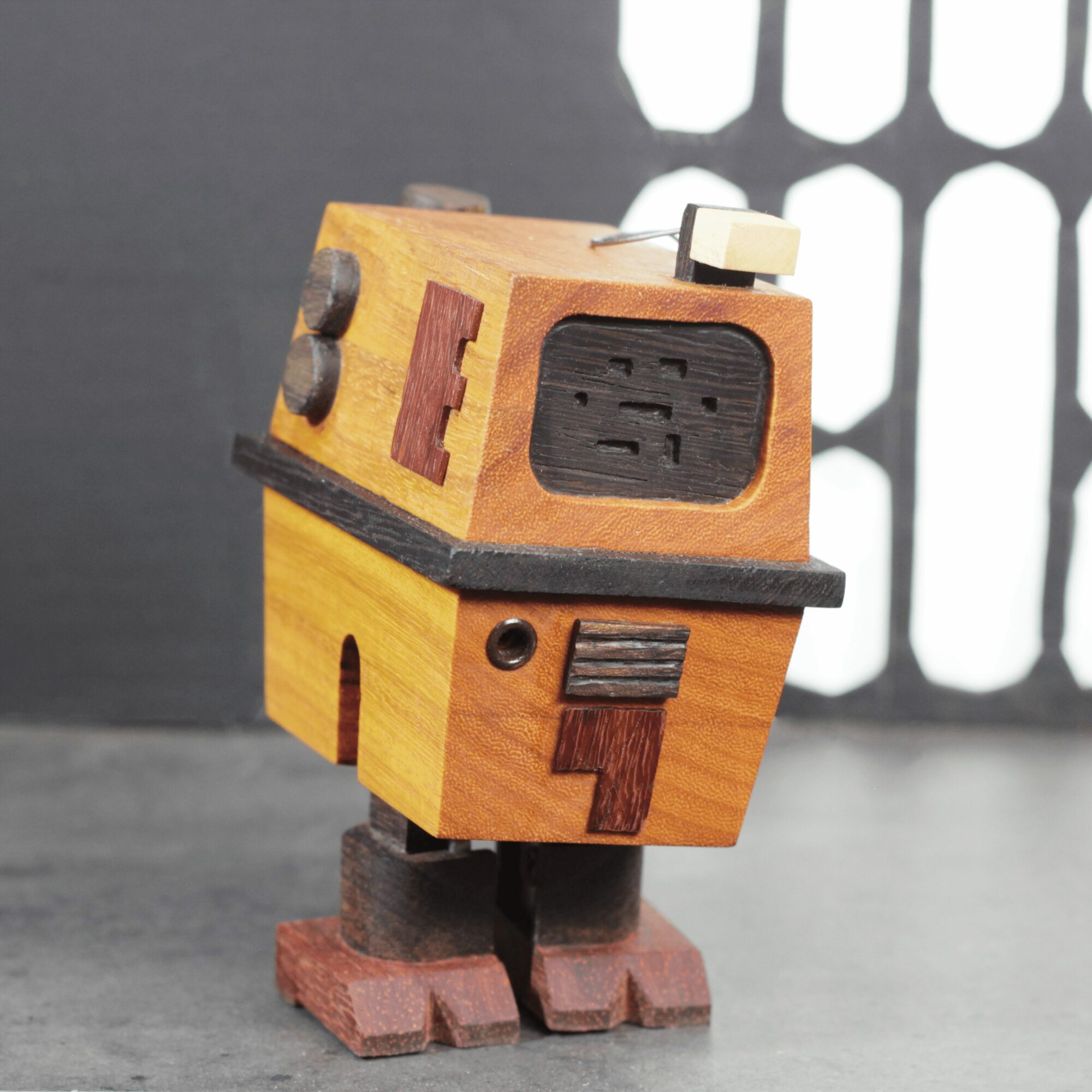 Wooden robot figure with a boxy body, rectangular head, and painted details, standing on a surface with a patterned background.