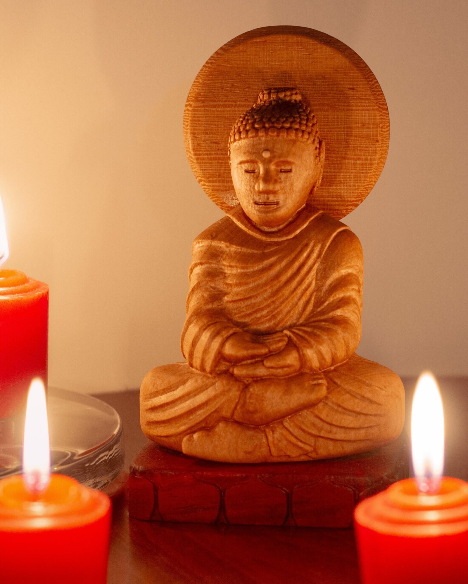 Wooden Buddha statue with crossed arms, seated on a base, surrounded by candles, against a plain background.
