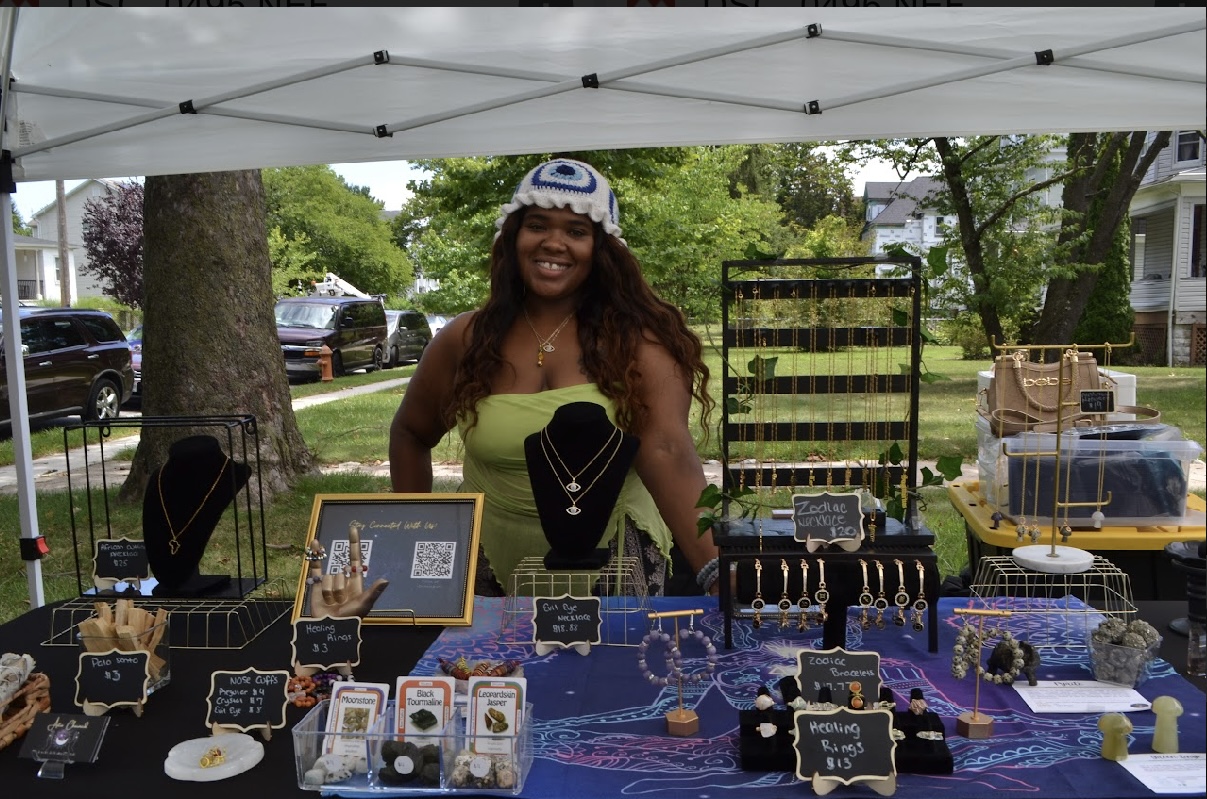 Woman smiling behind jewelry display at outdoor market stall with trees and houses in background.