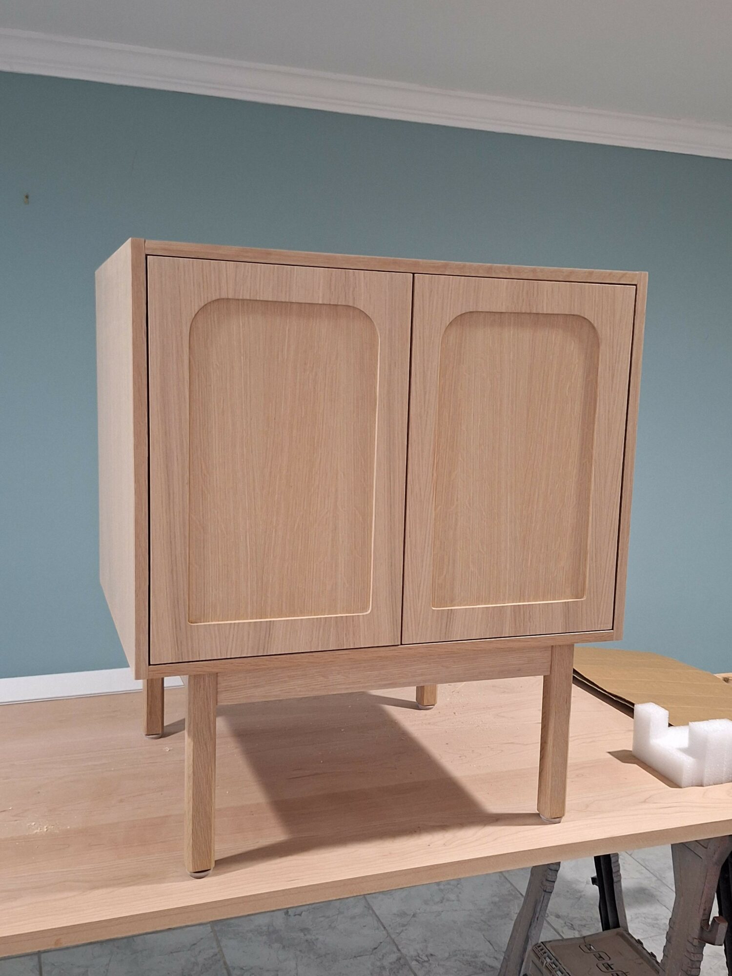 Wooden cabinet with two doors and a small open shelf, placed on a table against a blue wall.