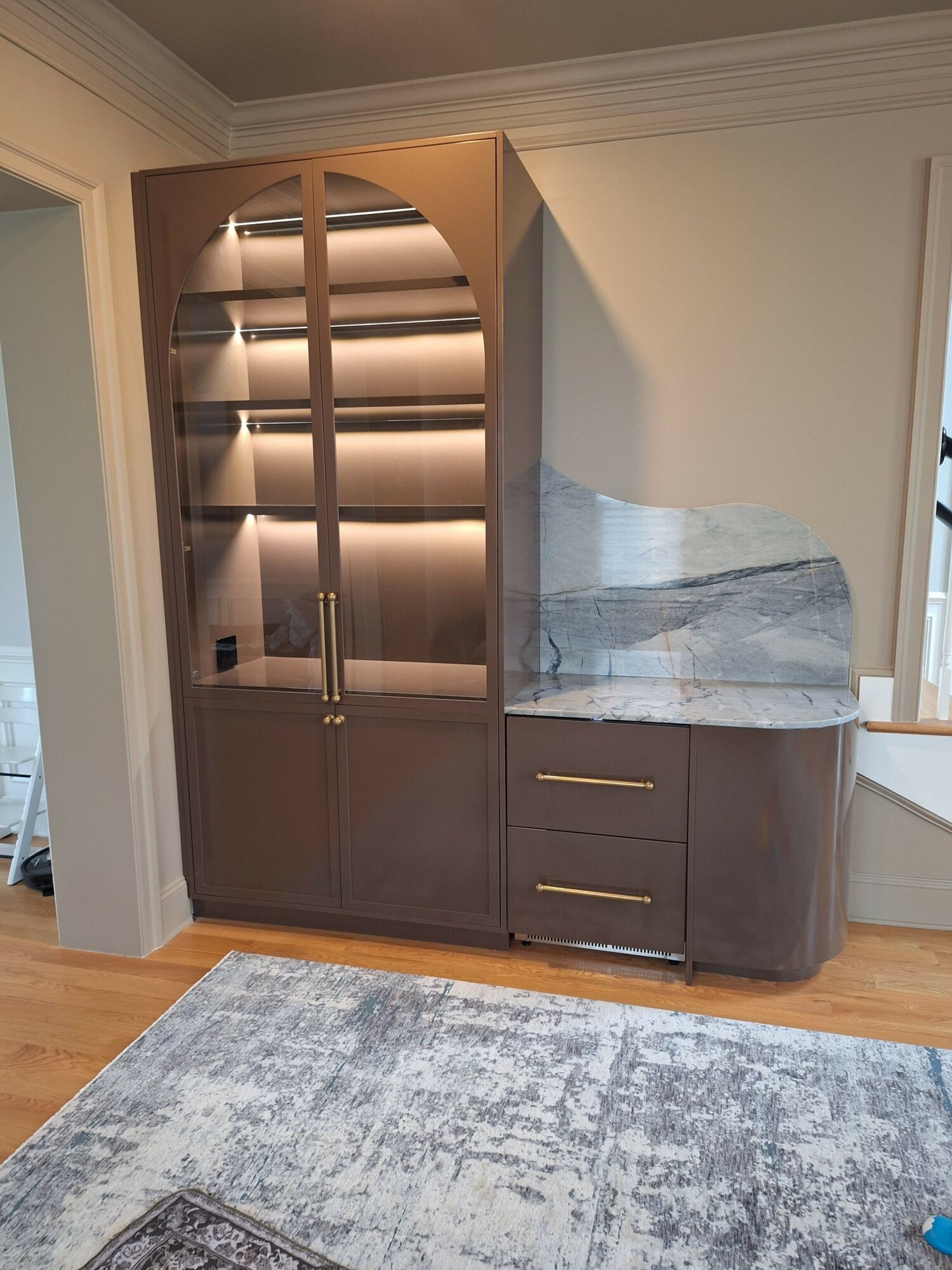 Brown cabinet with glass doors and a marble top, placed on a wooden floor next to a blue rug.