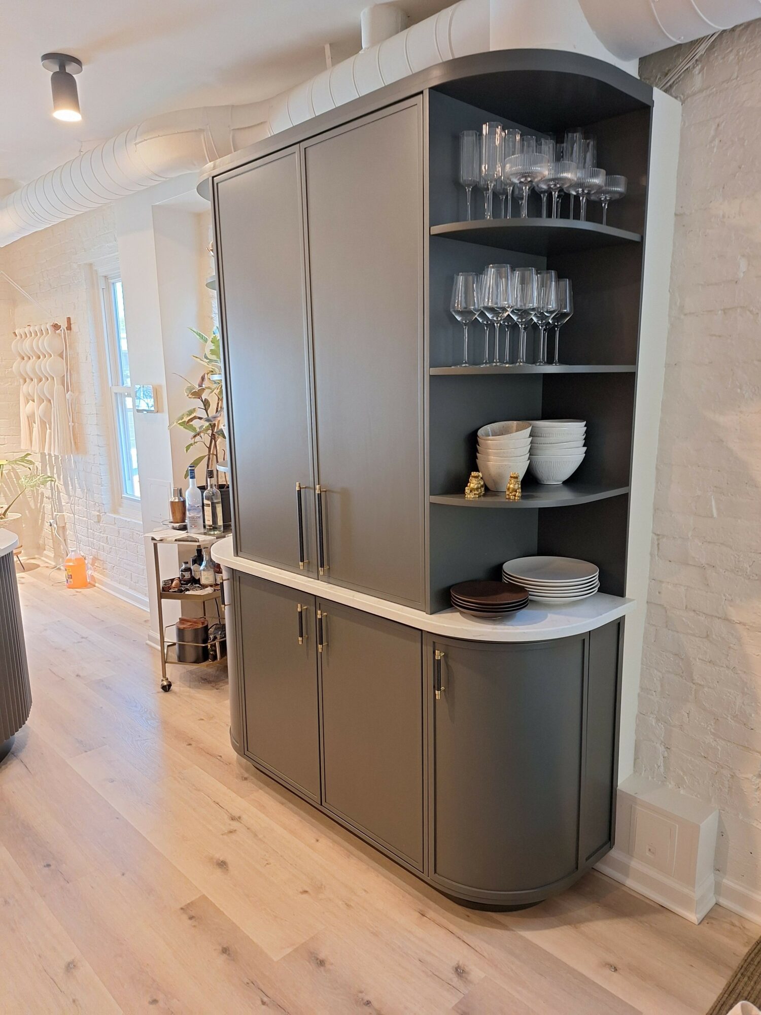 Kitchen cabinet with glassware, plates, and a small appliance on top, against a white brick wall and wooden floor.