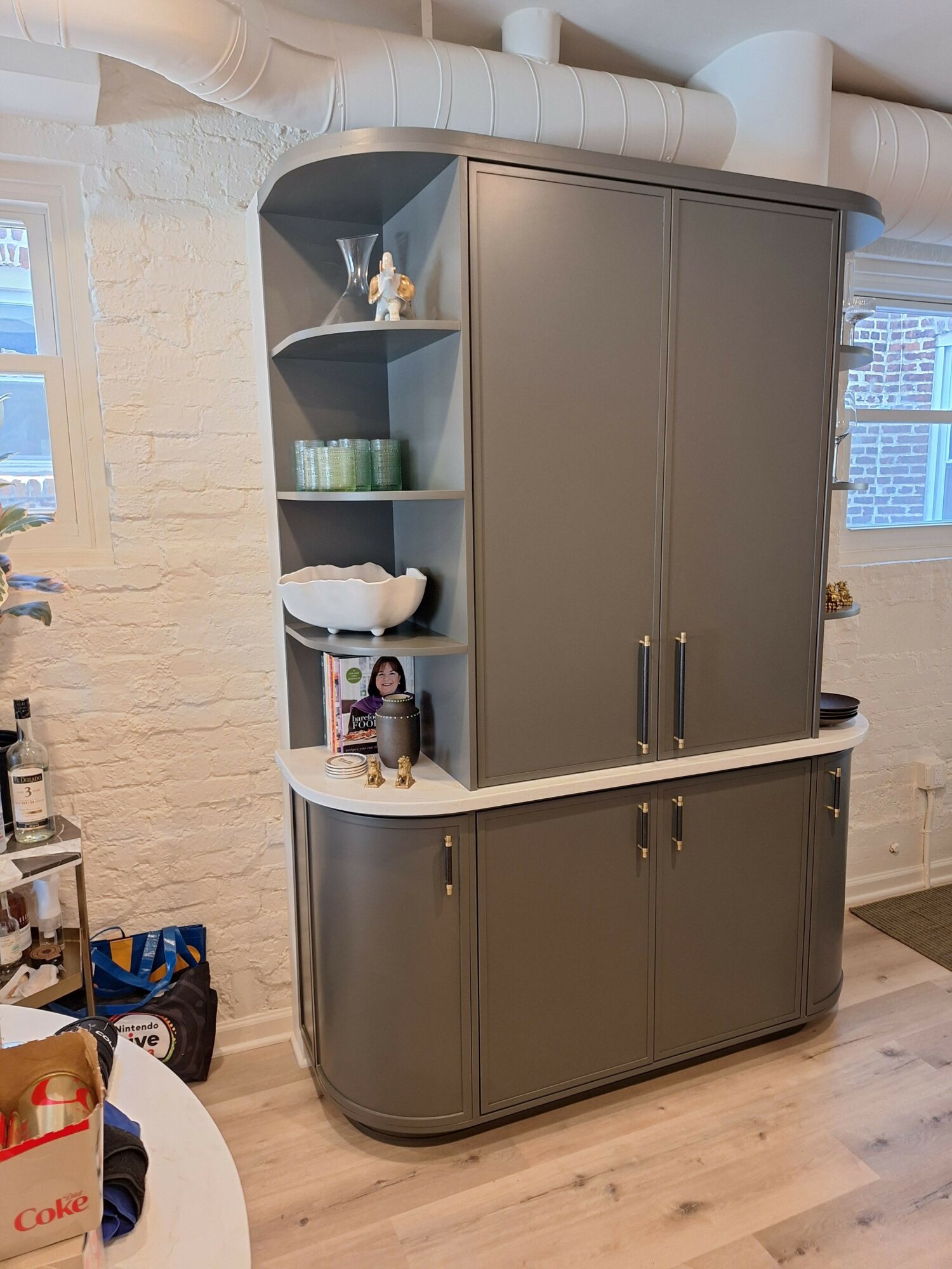 Gray storage cabinet with open shelves and closed compartments, placed on a wooden floor in a room with a brick wall.