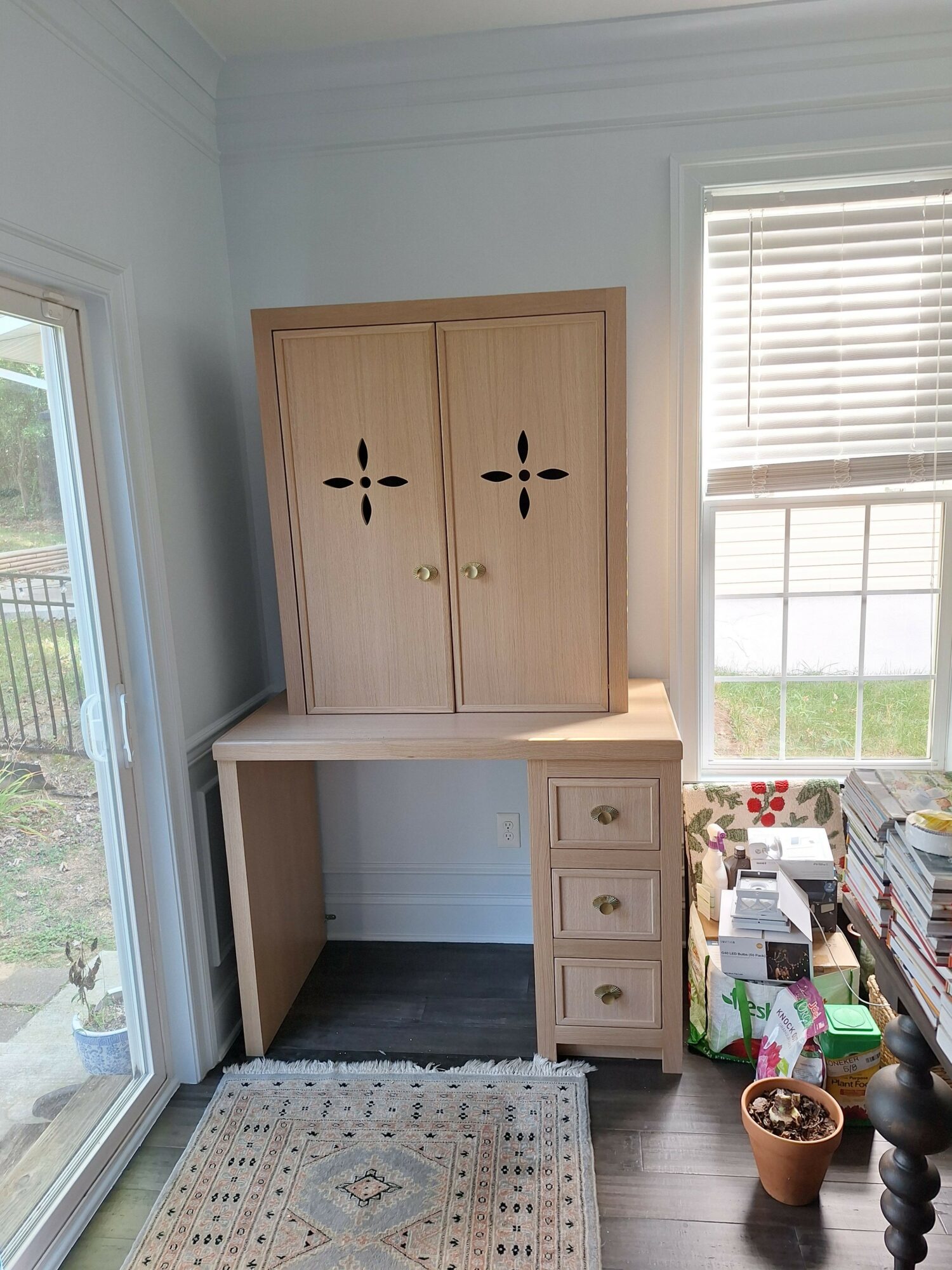 Wooden cabinet with open shelves and drawers, next to a window with blinds, in a room with a patterned rug.