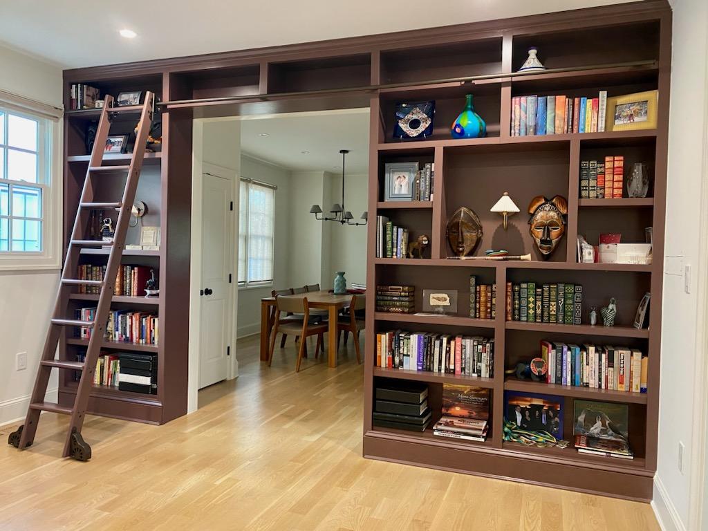 Living room with large wooden bookshelf, ladder, and view into dining area with table and chairs.