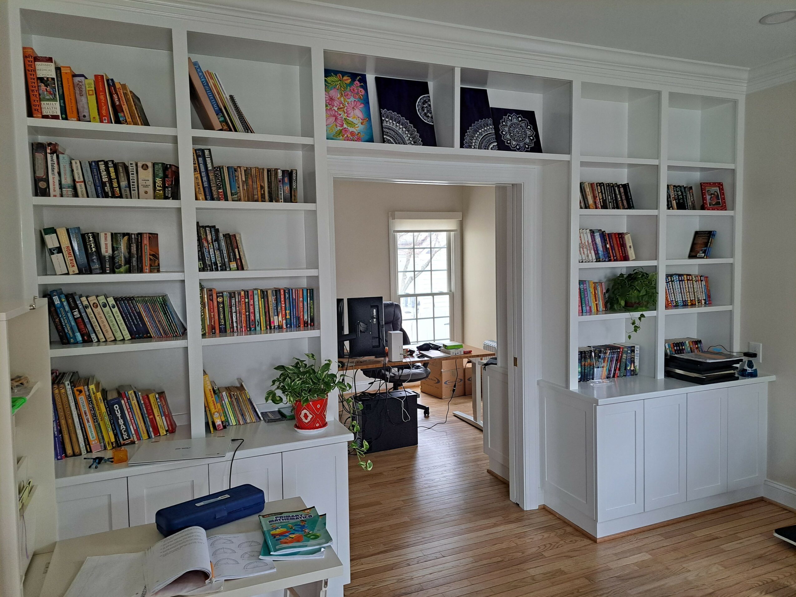 Bookshelves filled with books and decorative items, doorway leading to a room with a desk and window, wooden floor.