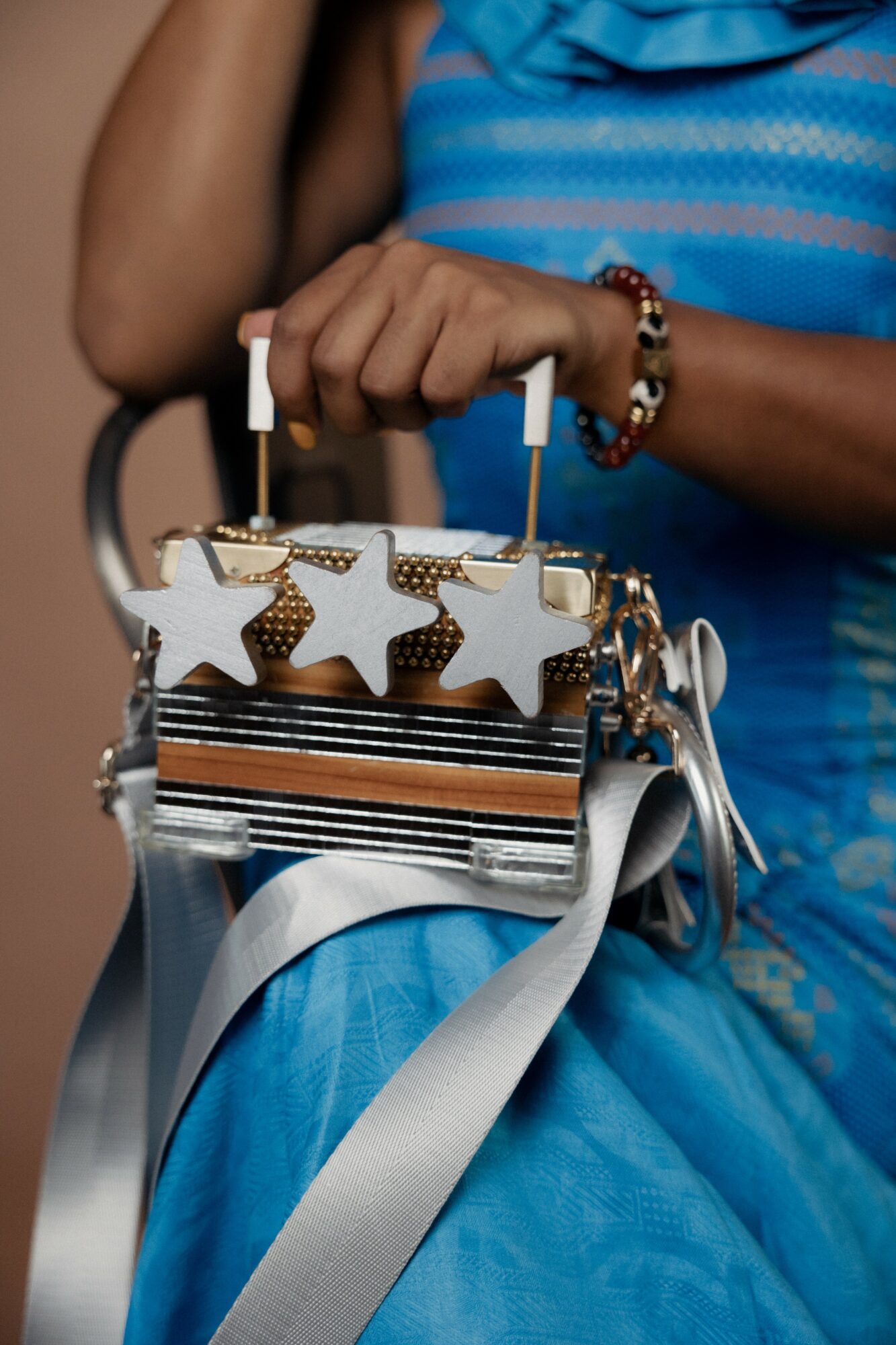 Person holding a silver handbag with star-shaped decorations, wearing a blue dress and a beaded bracelet.