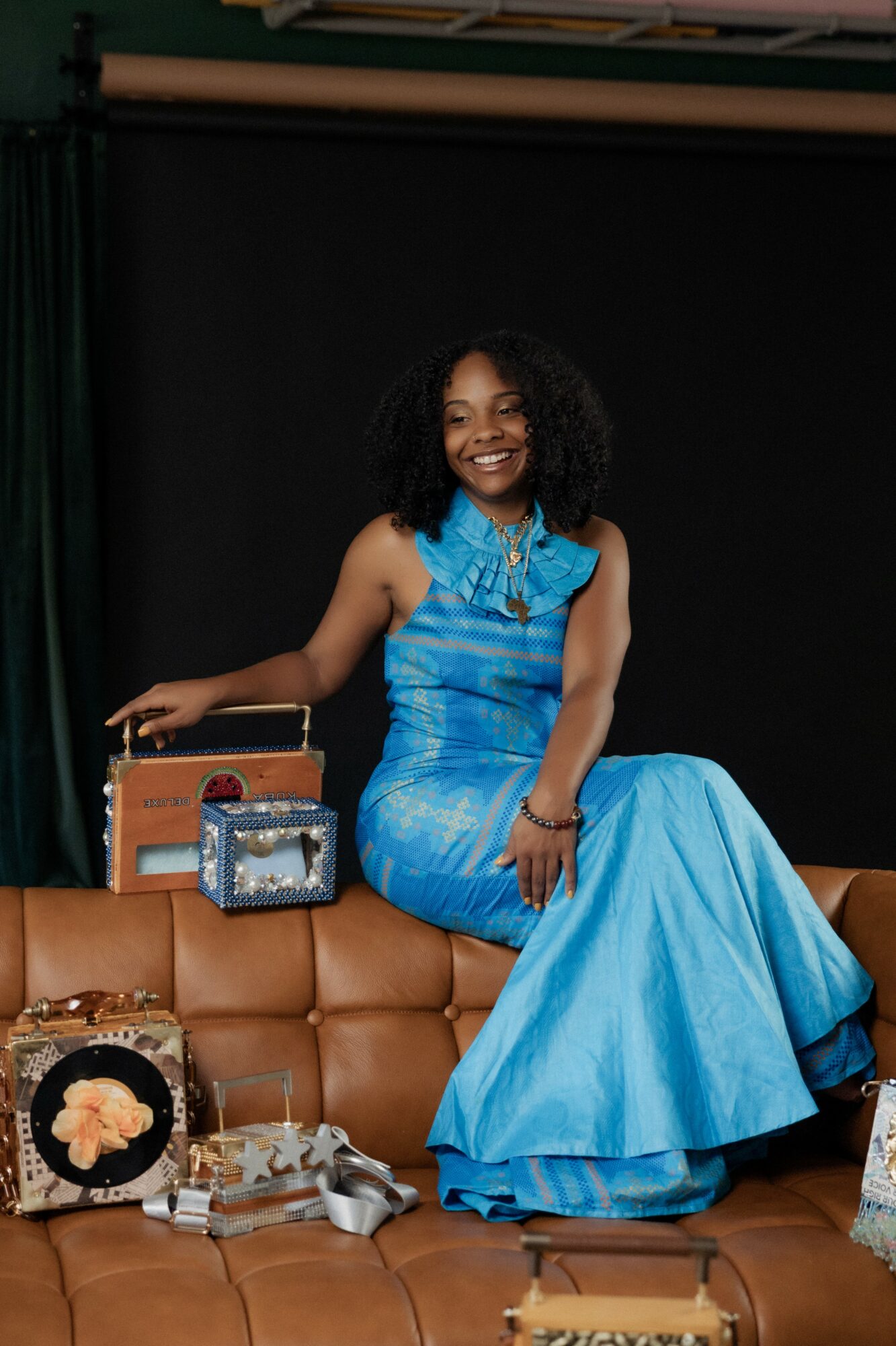 Woman in a blue dress sitting on a brown couch, smiling, with a black background.