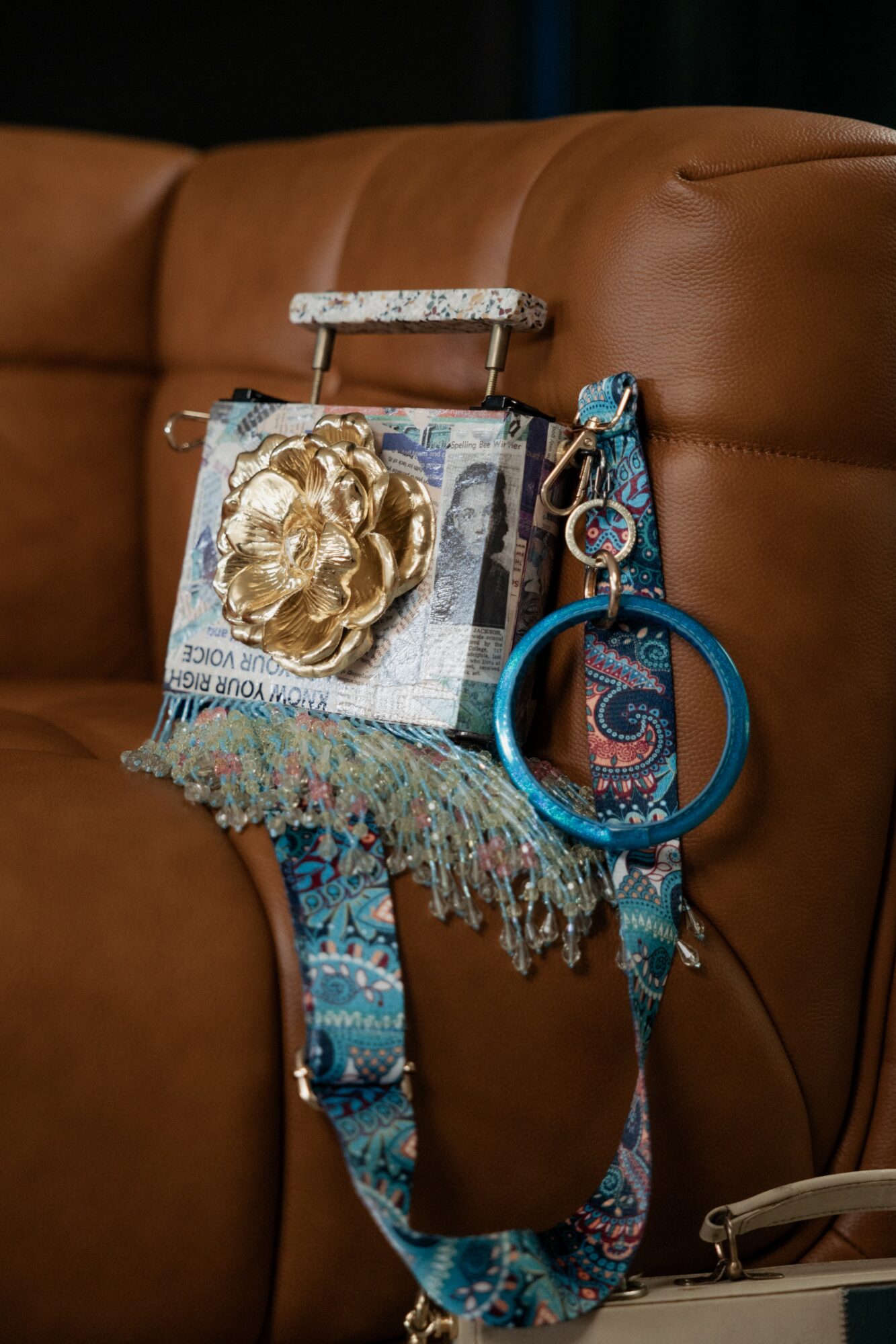 Decorative handbag with a large gold flower, hanging on a brown leather sofa, with a blue circular keychain and fabric strap.