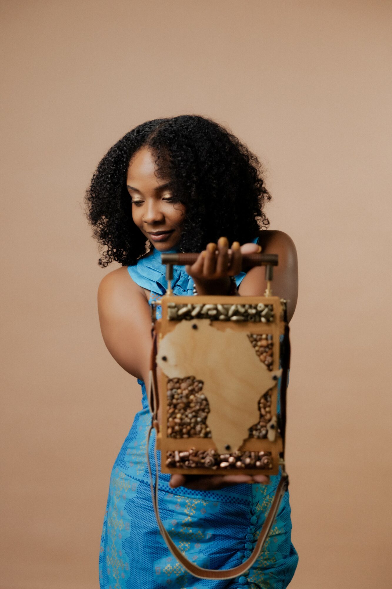 Young girl with curly hair in a blue dress holds a decorated box against a beige background.