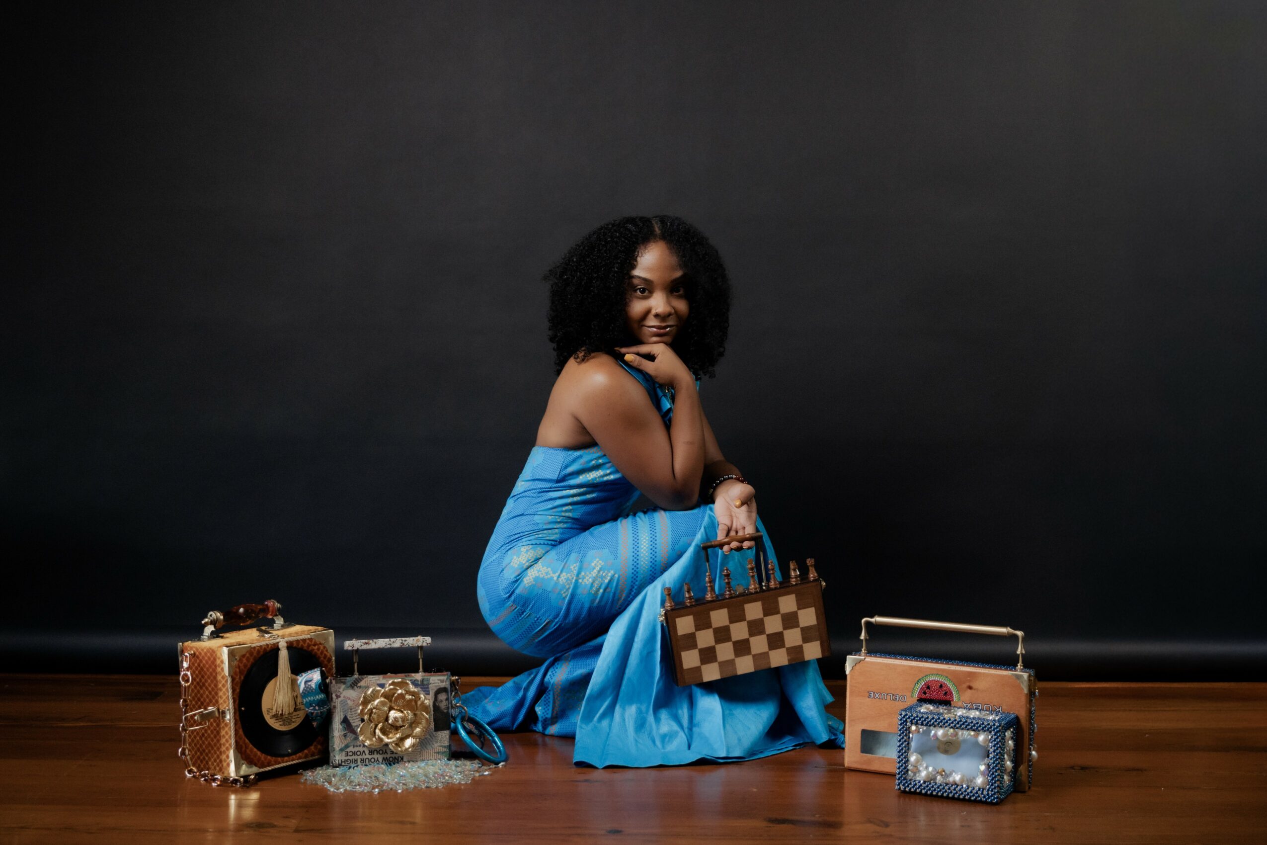 Woman in a blue dress sitting on the floor with various handbags and accessories around her, against a dark background.