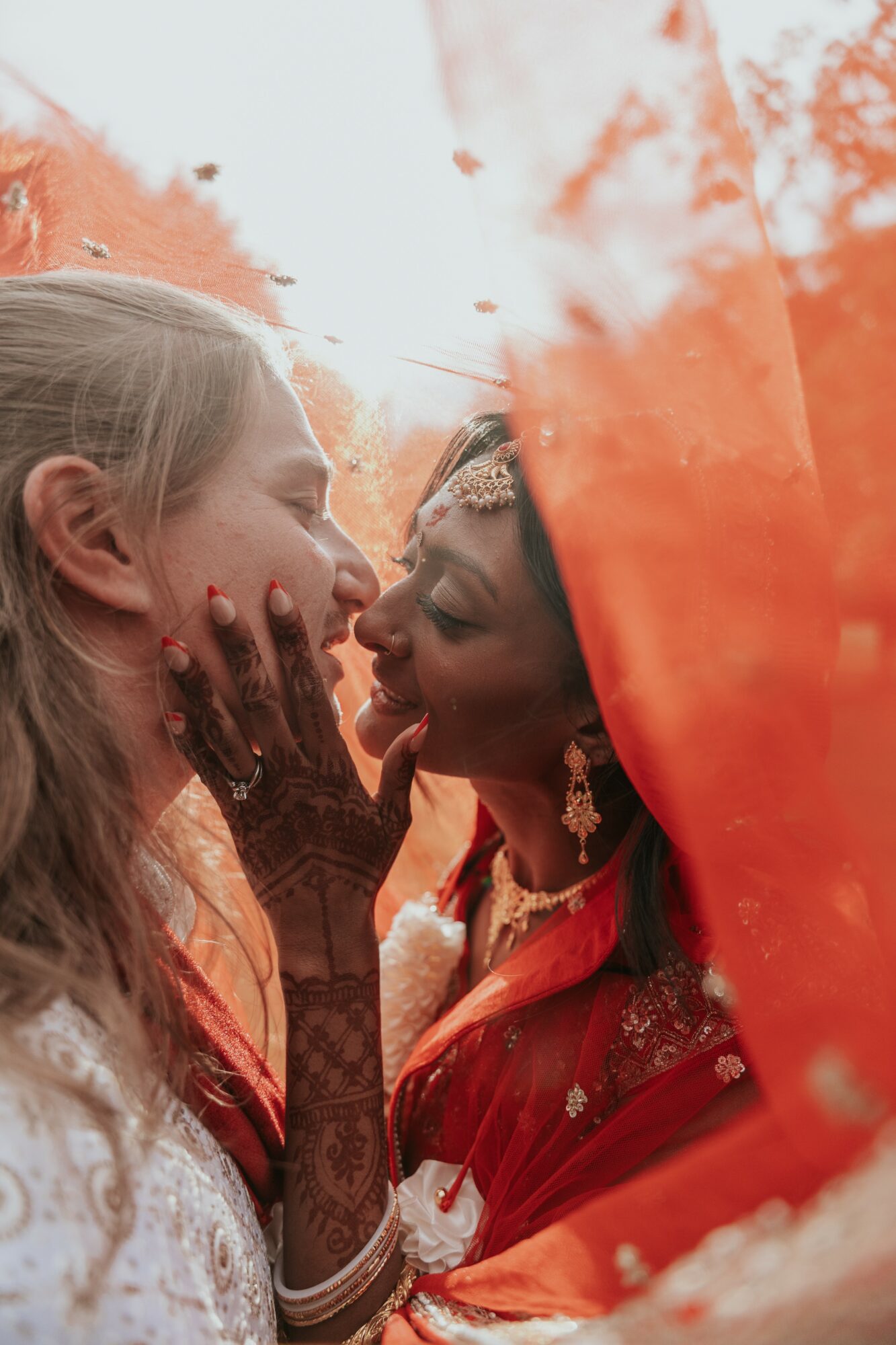 Two women face each other closely, touching noses, with one wearing traditional attire and jewelry, and the other with henna on her hand.