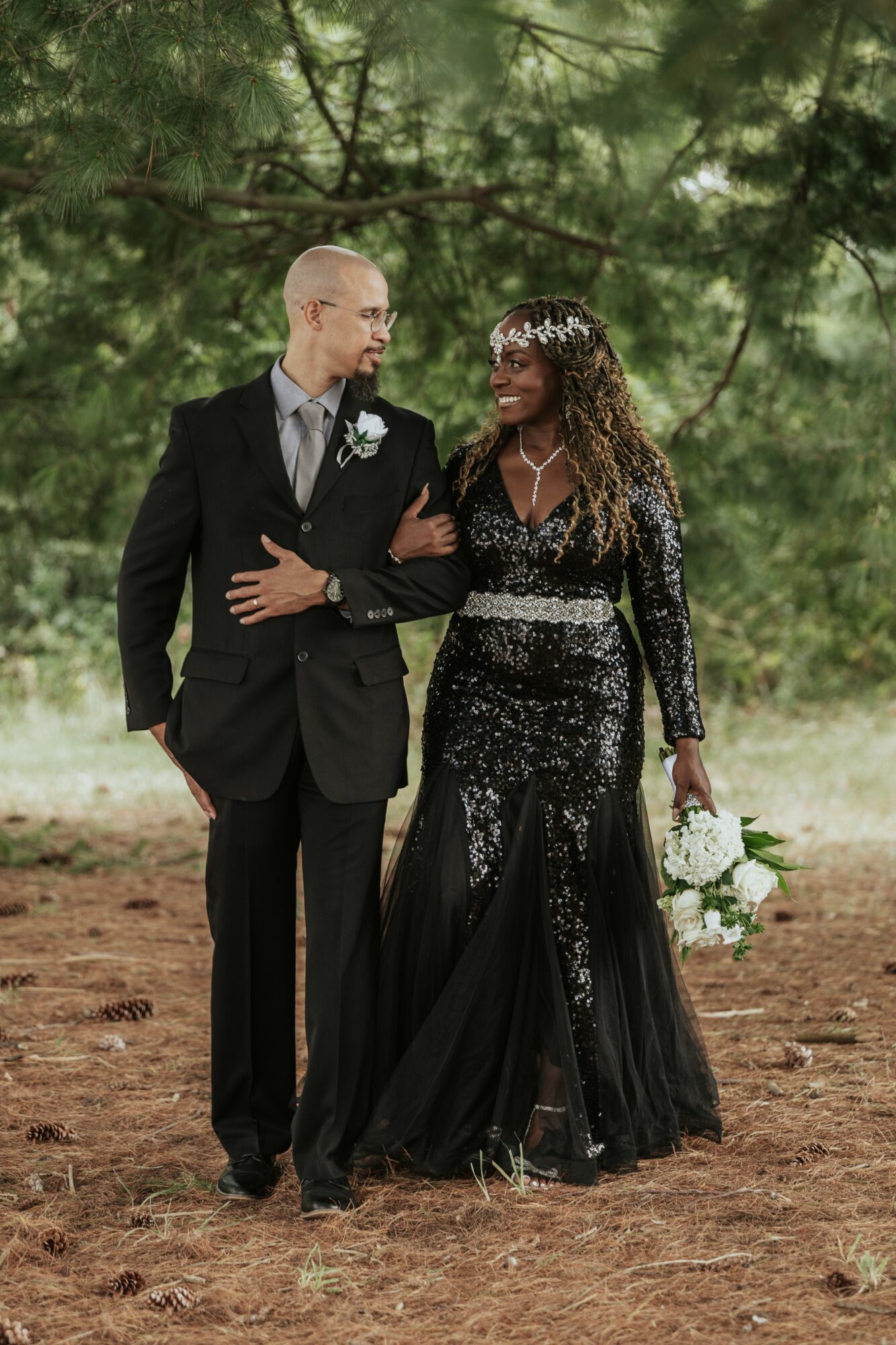 A couple walking outdoors on a dirt path surrounded by trees, dressed in formal attire, smiling at each other.
